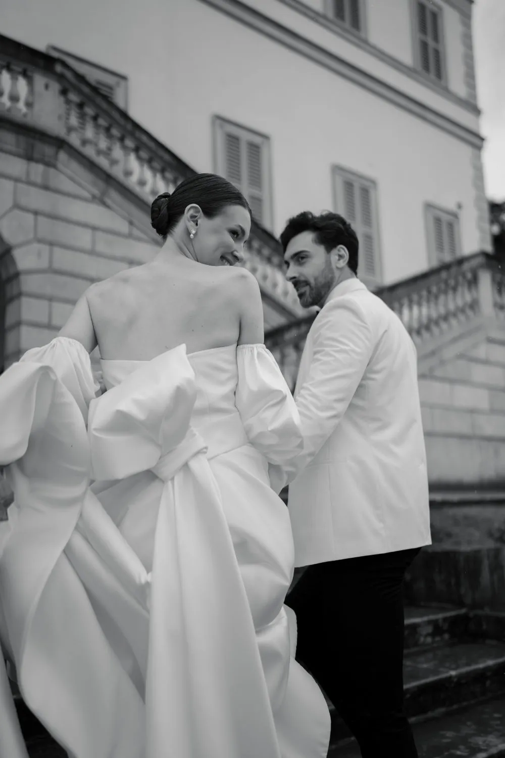 Bride in a bow-back gown and groom in a white blazer climbing stone steps by a villa