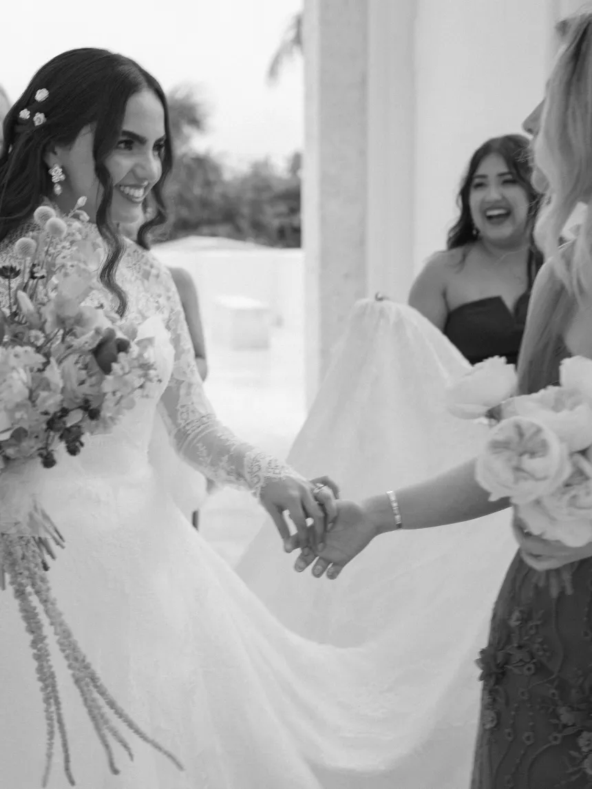 Bride in a lace gown laughing with guests as they carry her train, in black and white