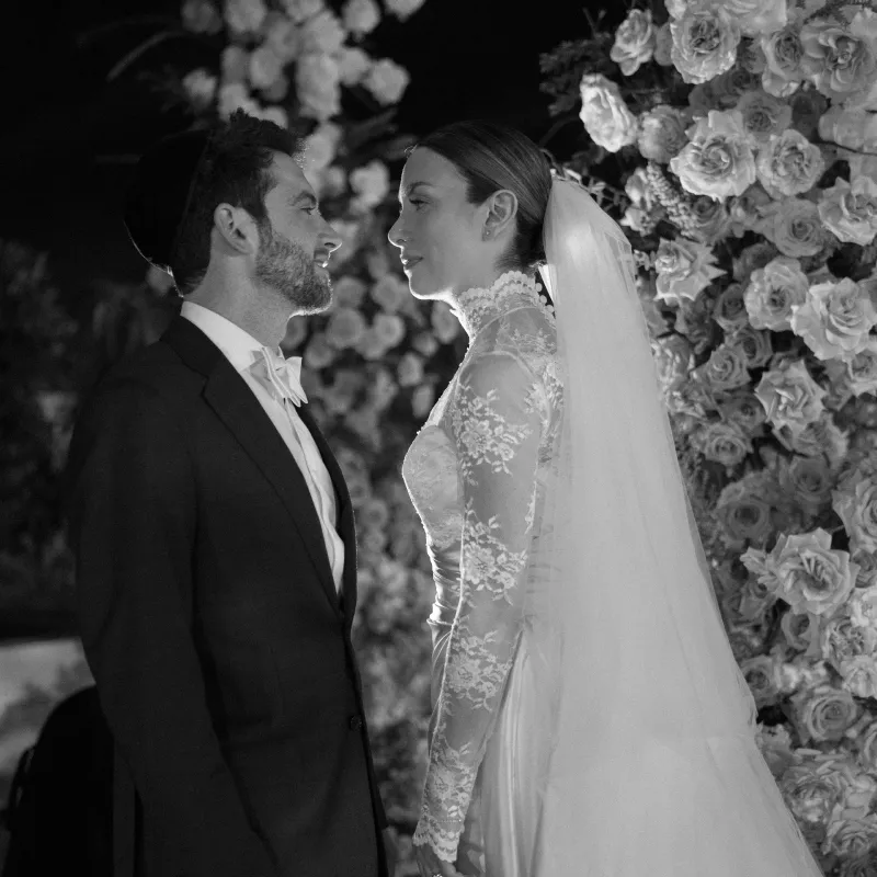 Bride in a lace gown and veil facing the groom in front of a wall of roses