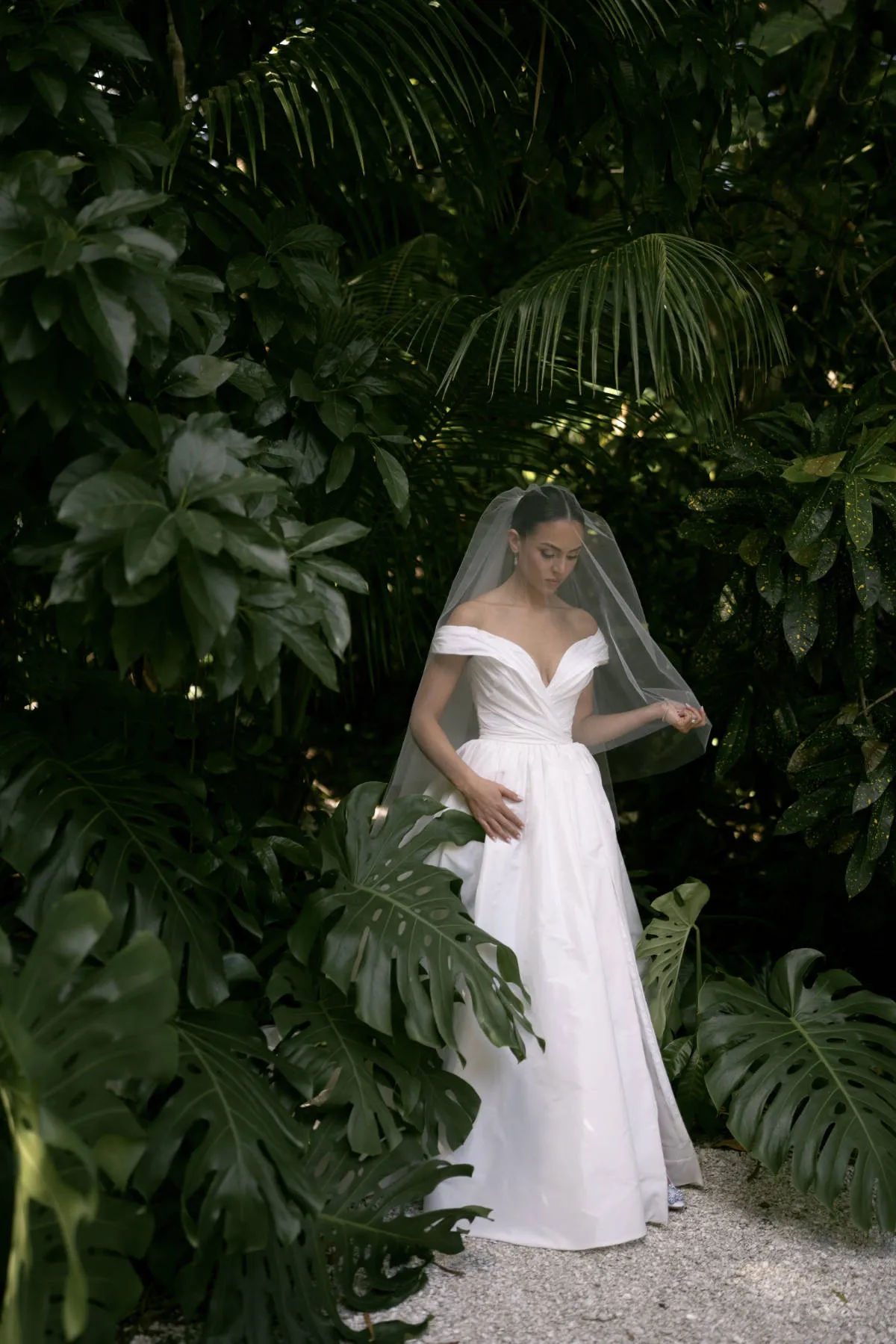 Bride in an off-shoulder gown and veil standing among tropical palms and leaves
