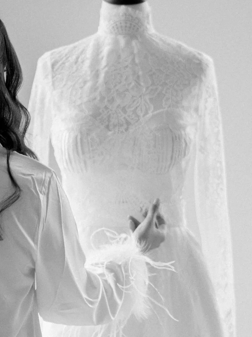 Bride in a silk robe reaching toward a high-neck lace gown on a dress form