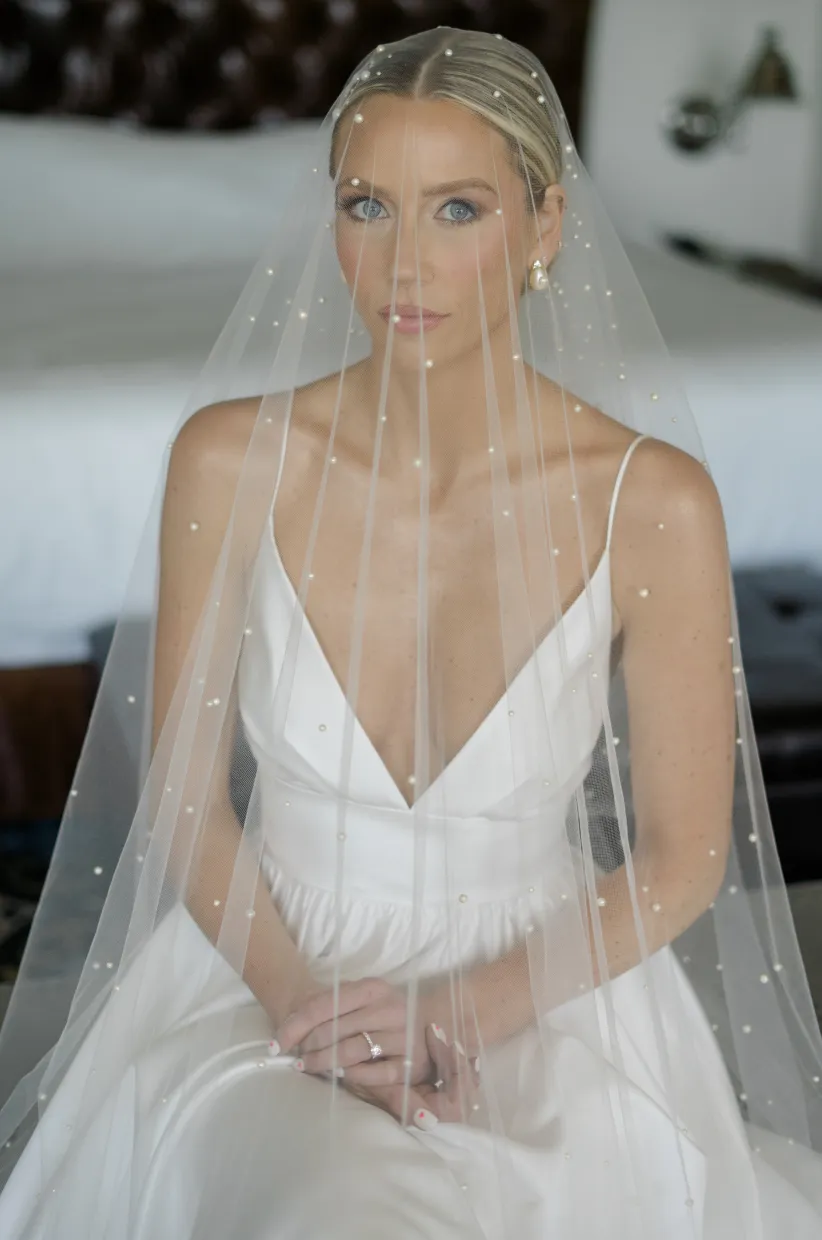 Bride seated in a slip gown under a pearl-scattered veil, looking at the camera