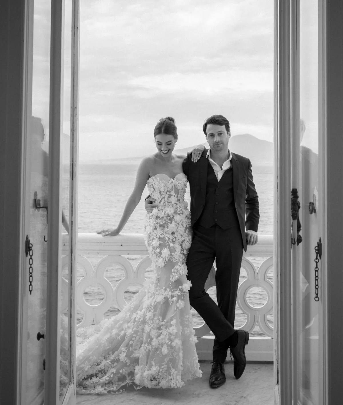 Bride and groom leaning on a balcony railing above the sea, seen through open doors