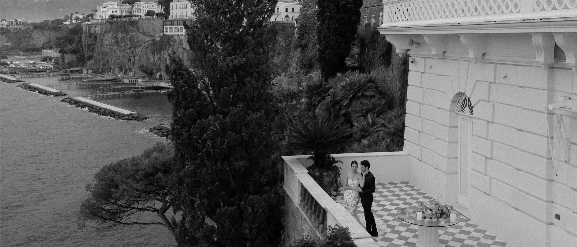 Distant view of a couple on a checkered villa terrace above a cliffside harbour