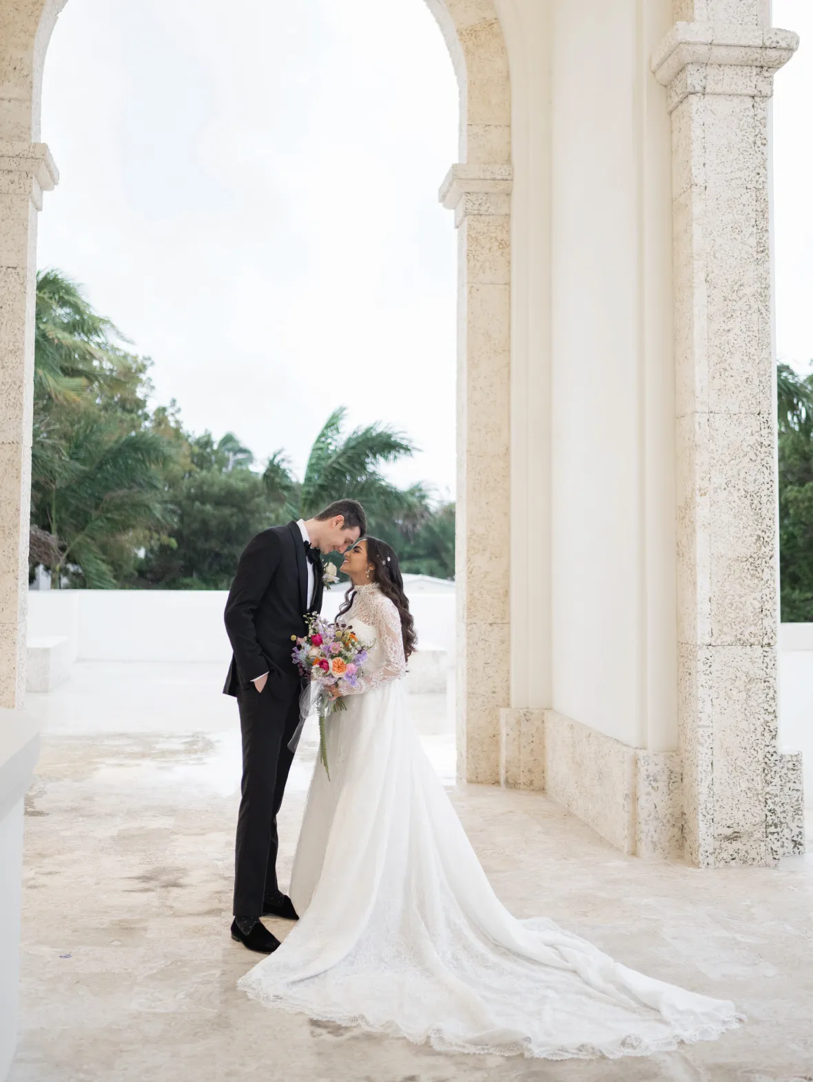 Bride with a bright bouquet and groom touching foreheads under a stone archway