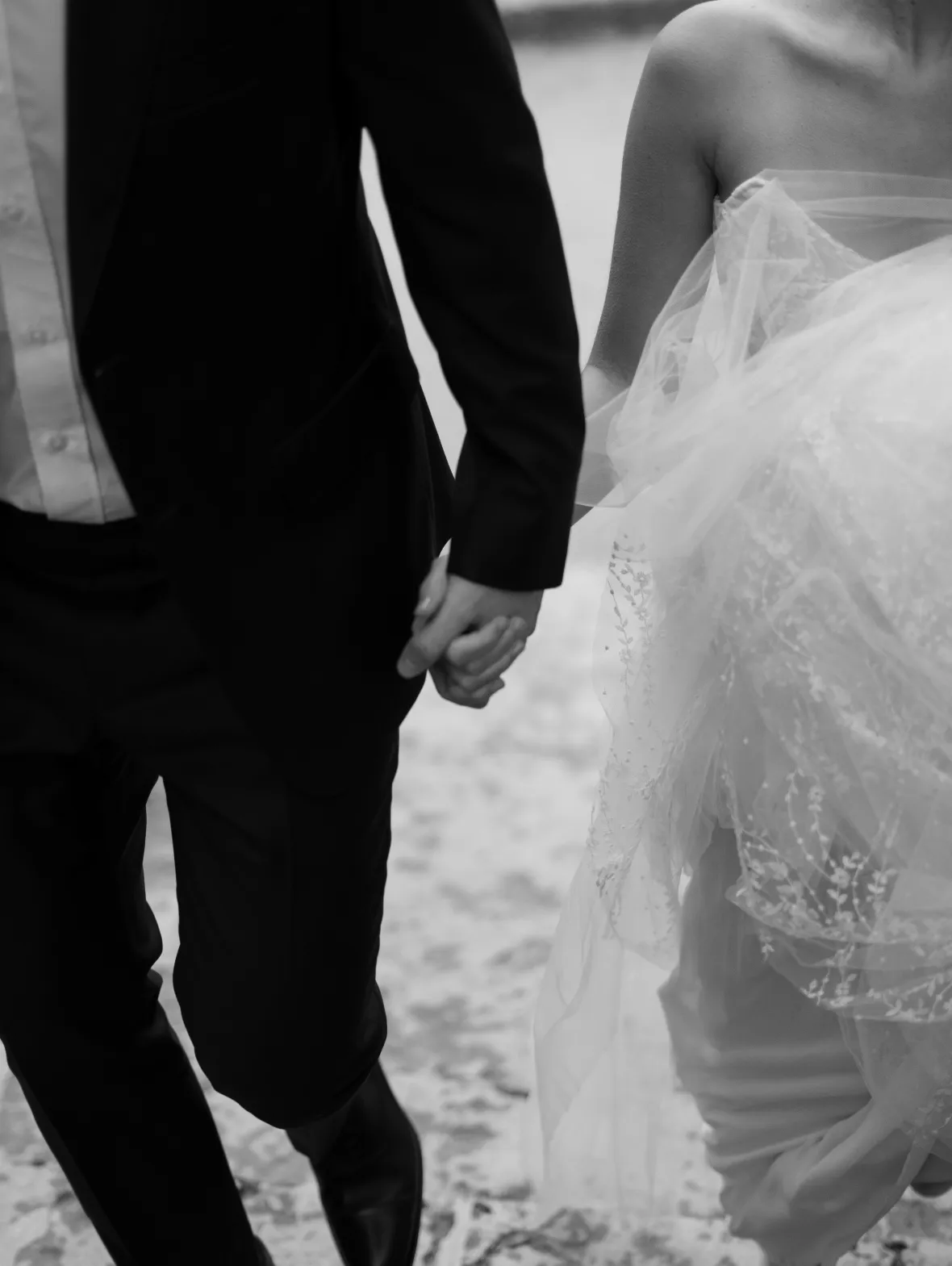 Bride and groom holding hands as they walk across sand, seen from behind