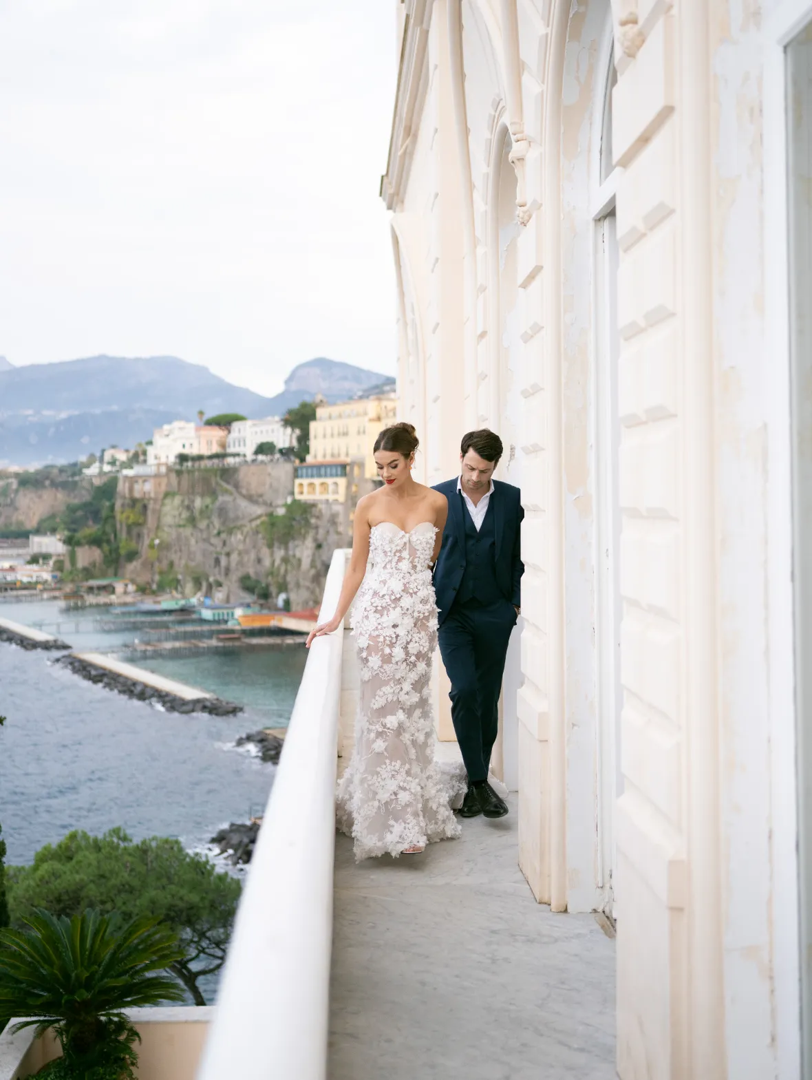 Bride and groom walking a narrow balcony above a cliffside harbour town