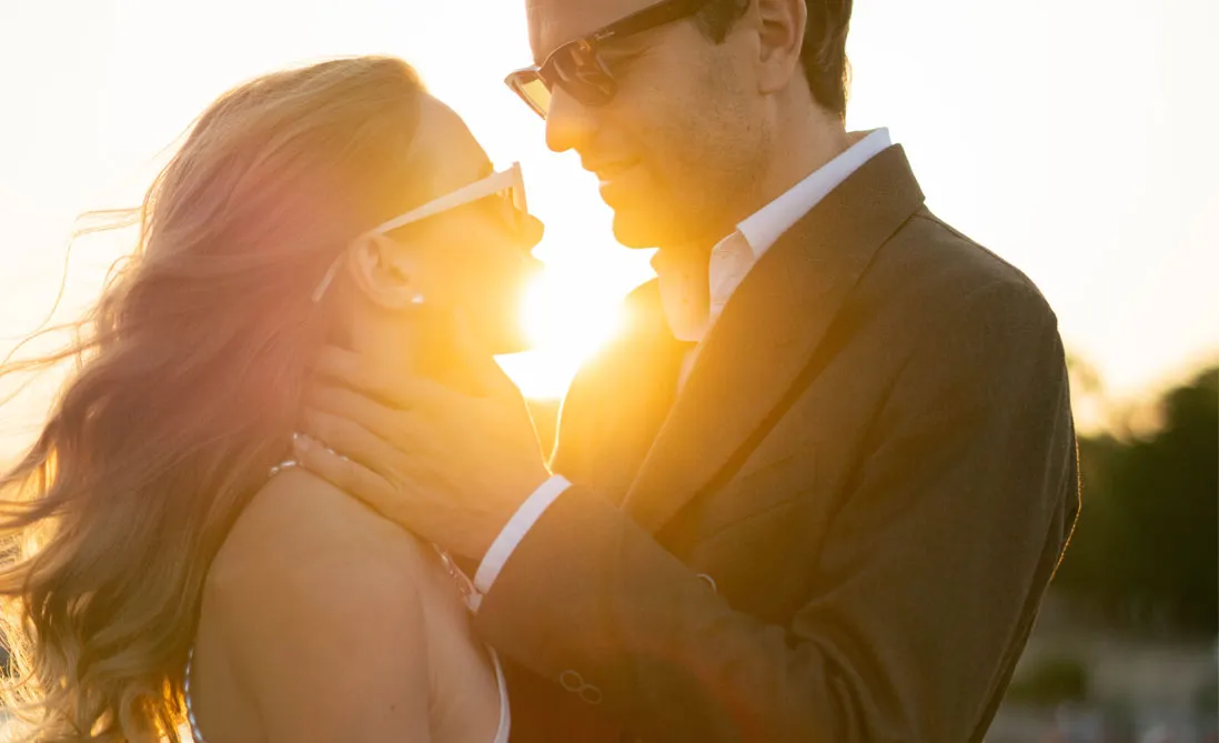 Couple in sunglasses leaning in close on a bridge, backlit by low evening sun