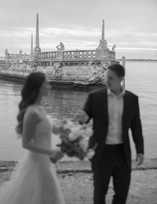 Bride with a bouquet and groom walking by the water, a carved stone barge behind them
