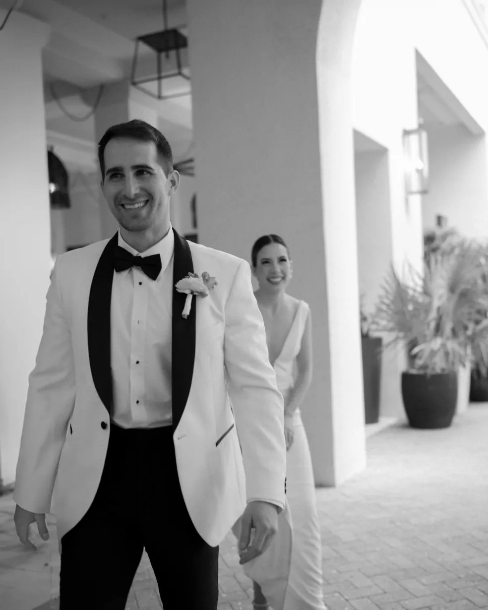 Groom in a white dinner jacket smiling as the bride approaches behind him