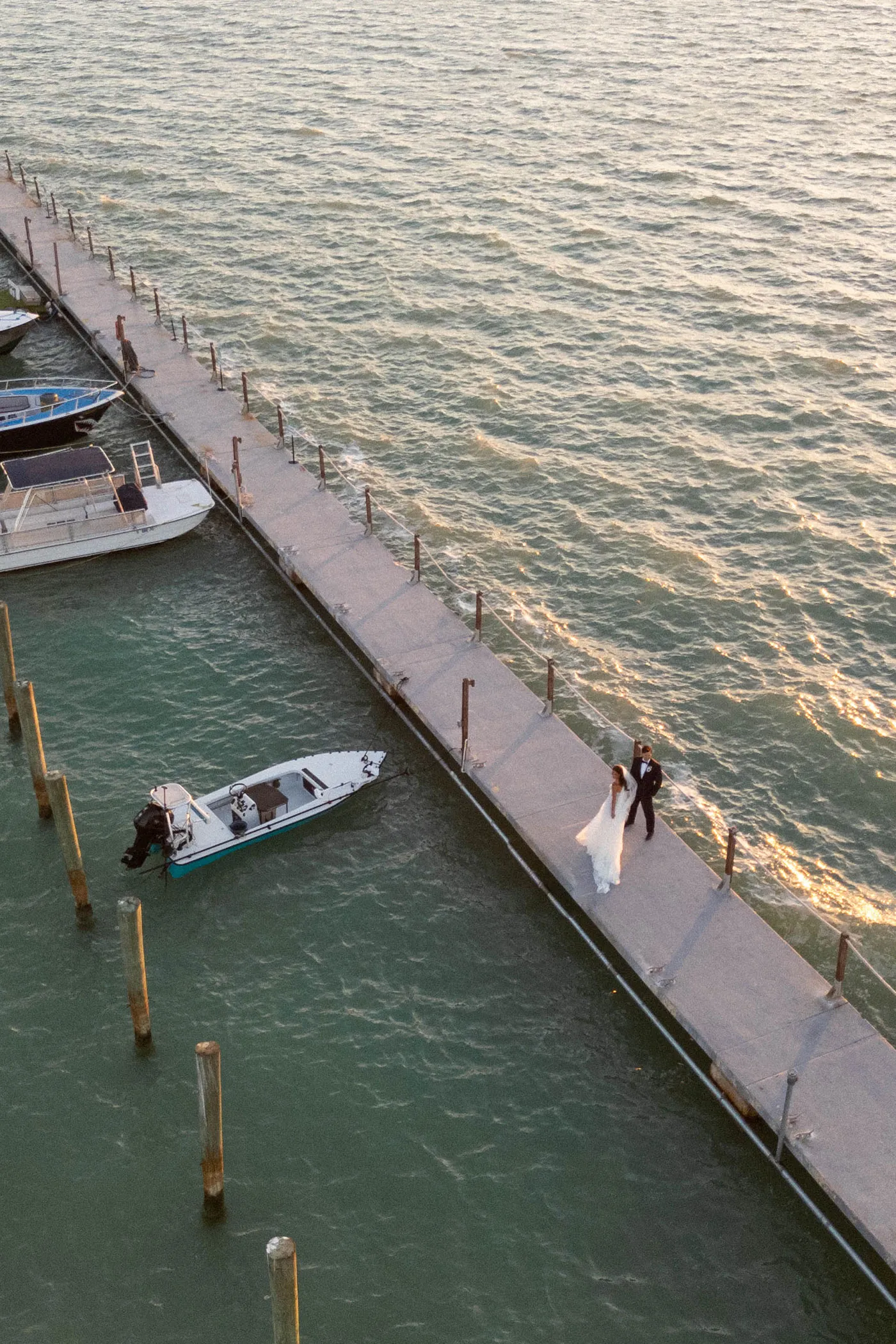 Bride and groom walking a long dock past moored boats at sunset, seen from above