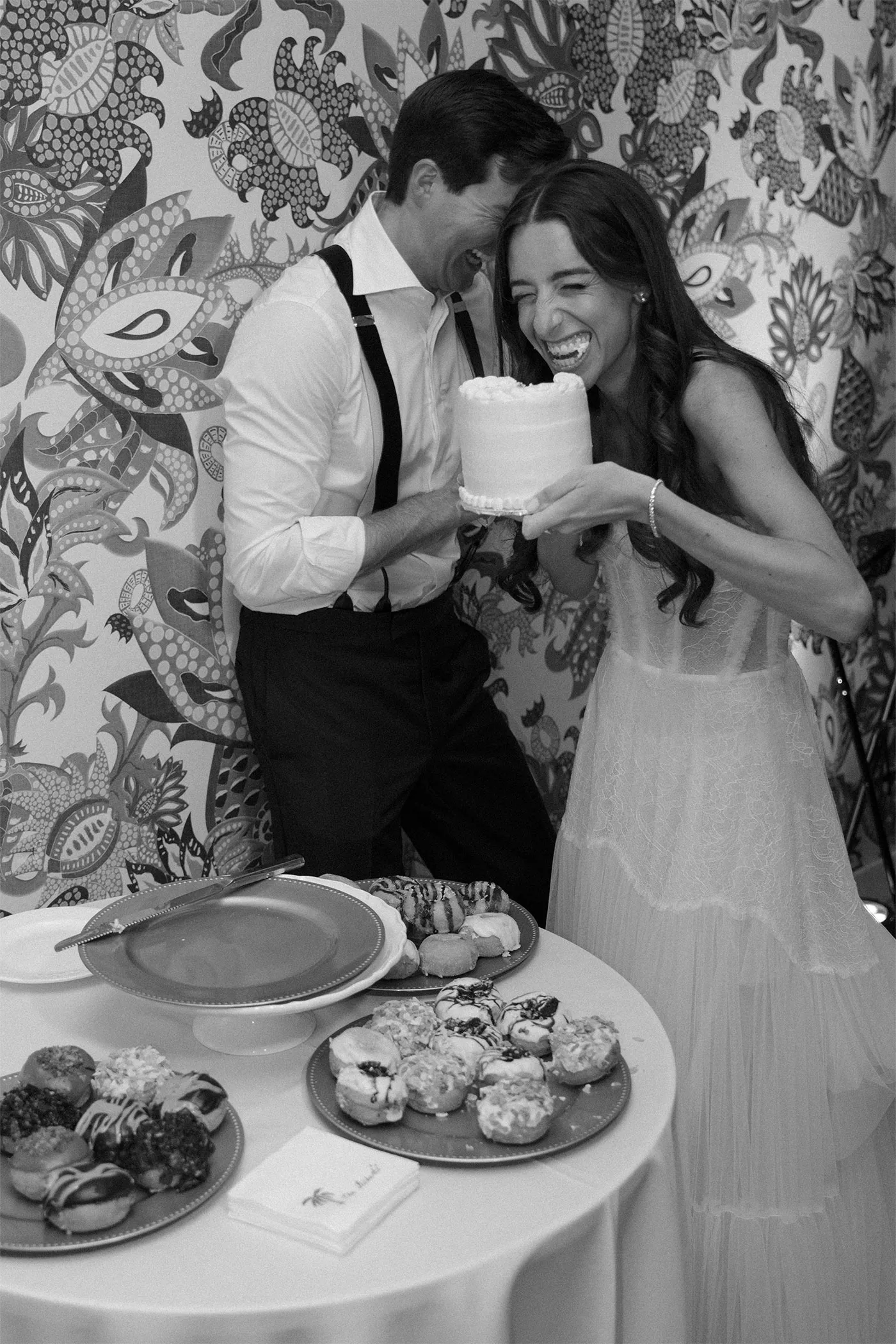 Bride laughing as she bites a small cake the groom holds, beside a doughnut table
