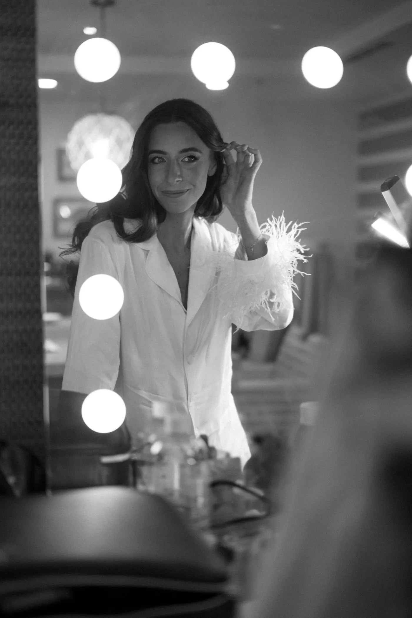 Bride in feather-trimmed pyjamas reflected in a lit vanity mirror