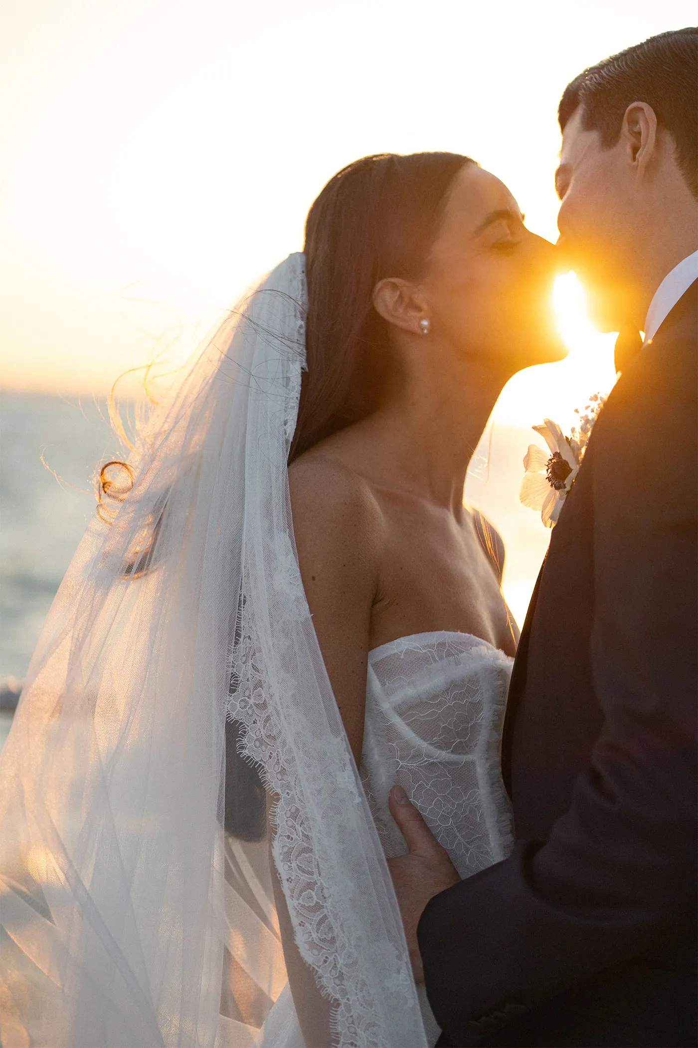 Bride in a lace veil and groom leaning in to kiss, backlit by the setting sun over water