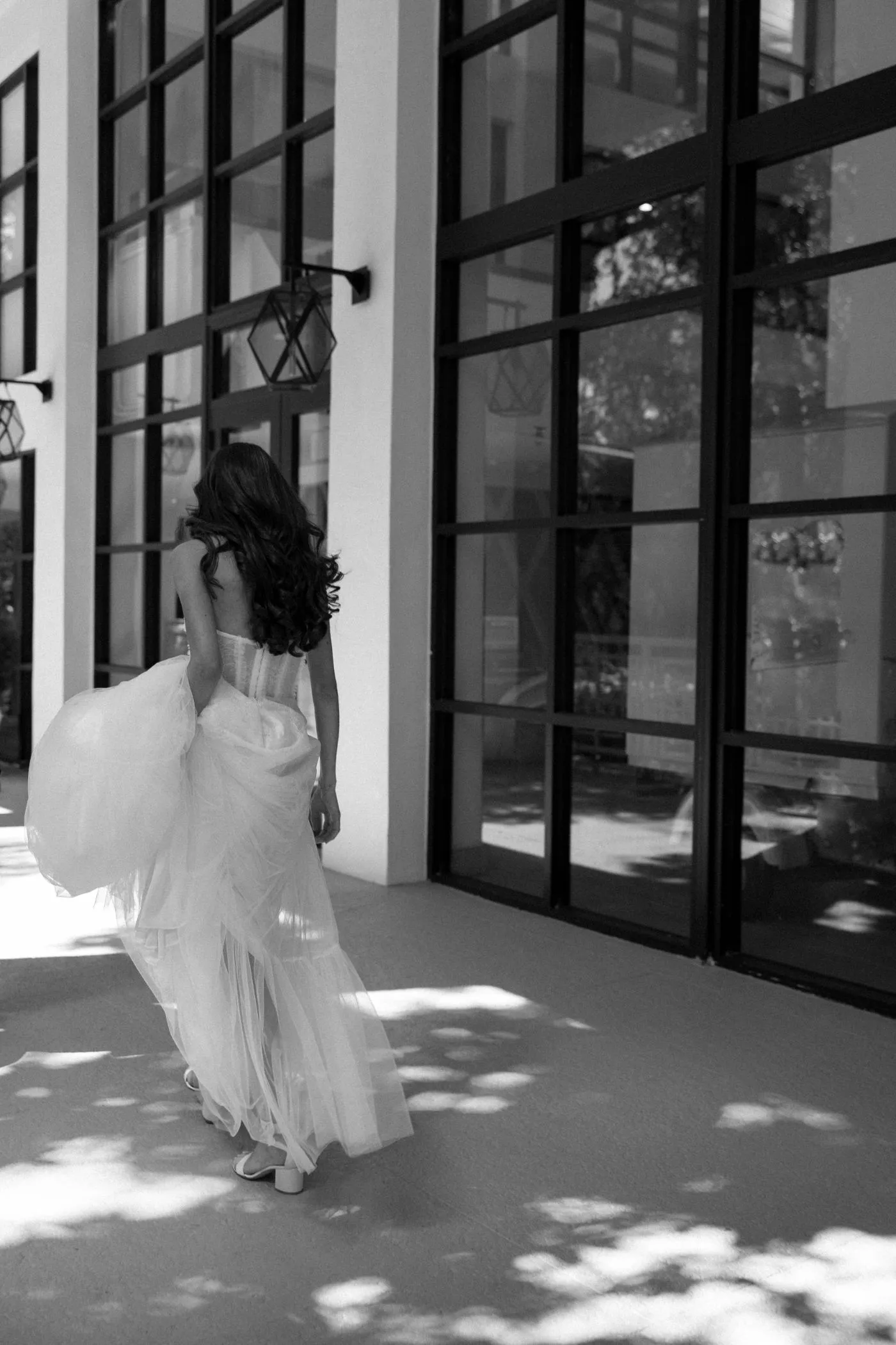 Bride from behind lifting her tulle skirt as she walks past tall windows