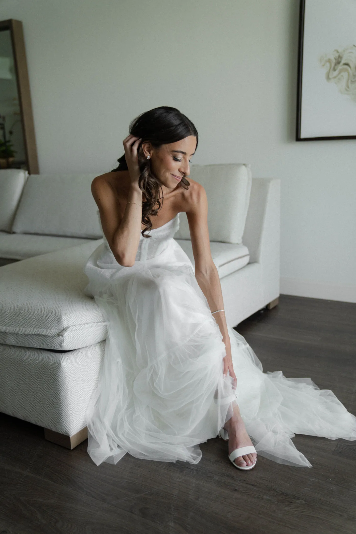 Bride in a strapless tulle gown seated on a white sofa, tucking back her hair