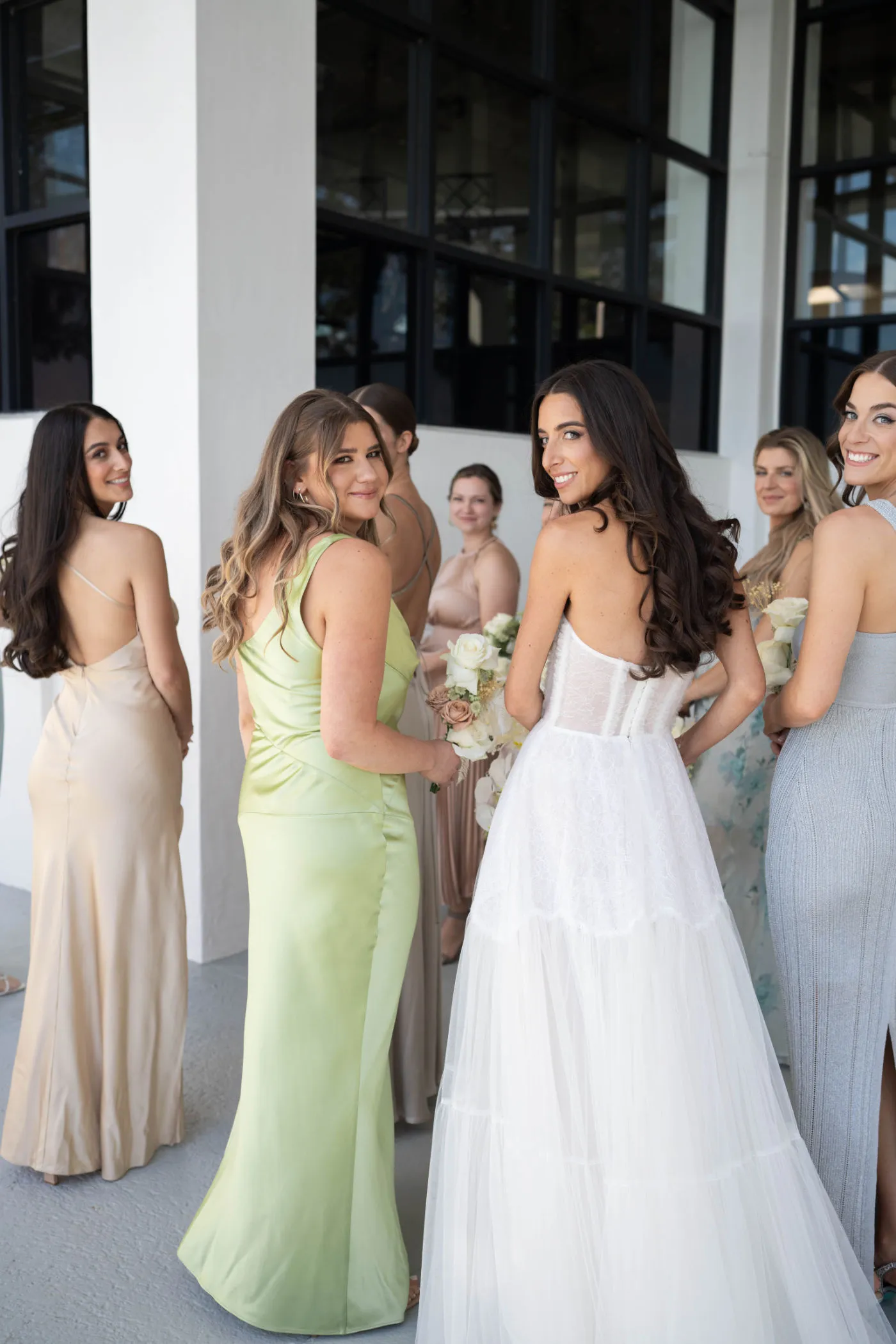 Bride and bridesmaids in pastel gowns looking back over their shoulders on a covered terrace