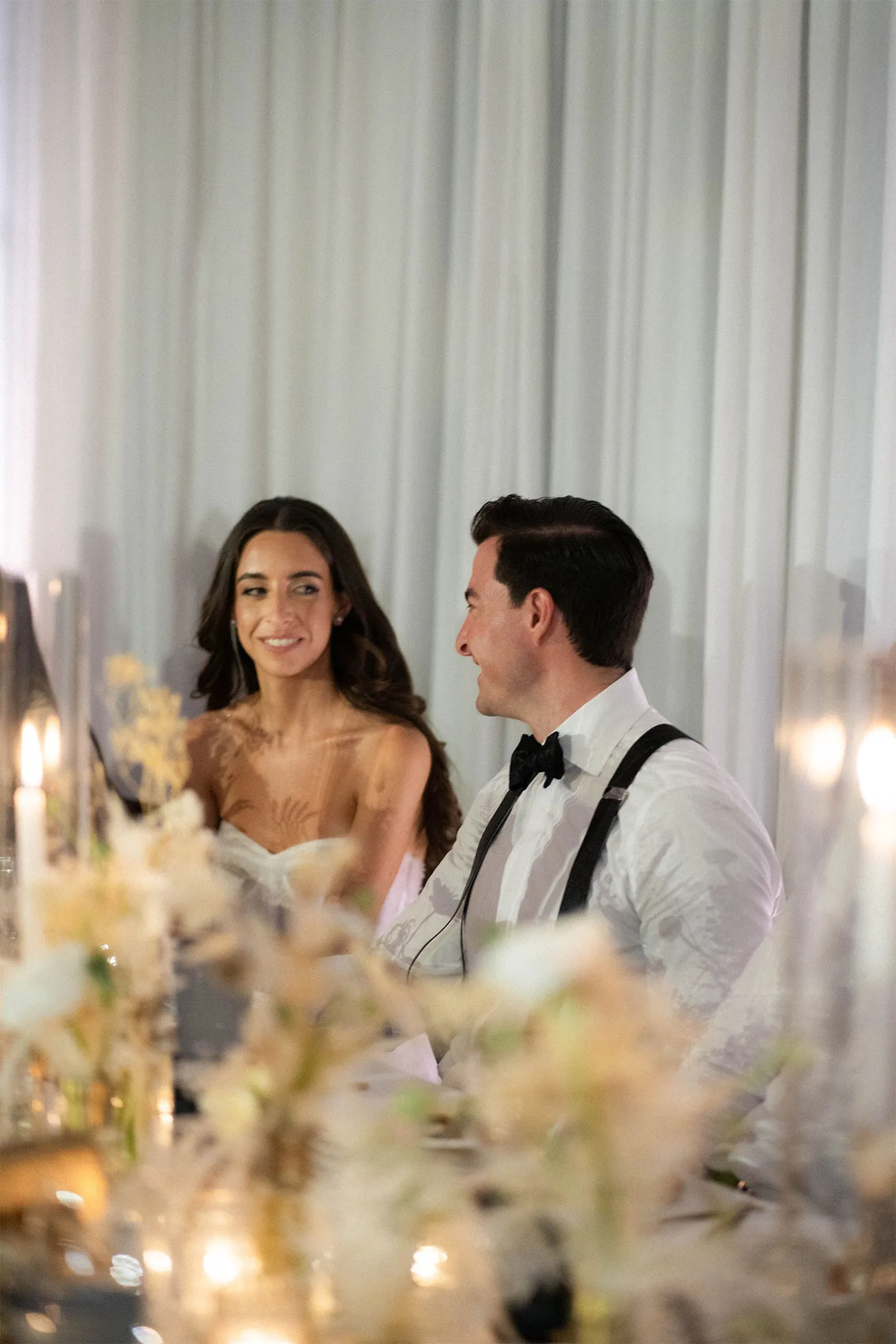 Bride and groom seated at a candlelit reception table, smiling at each other