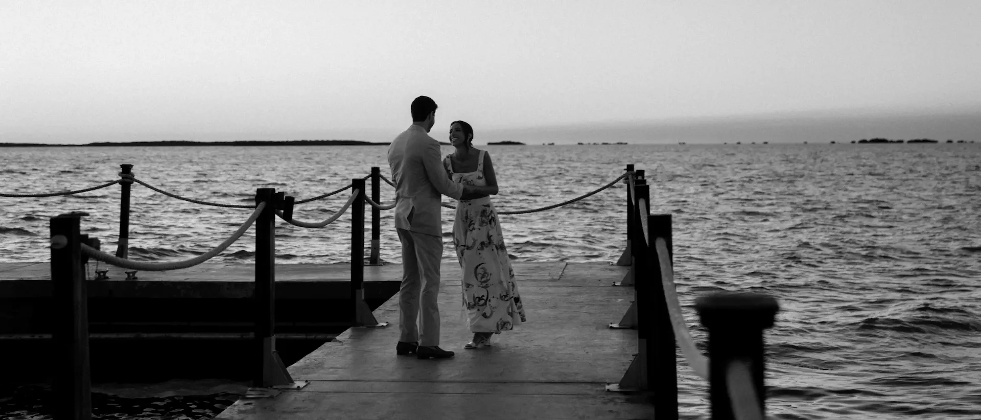 Couple standing at the end of a dock at dusk with open water behind them