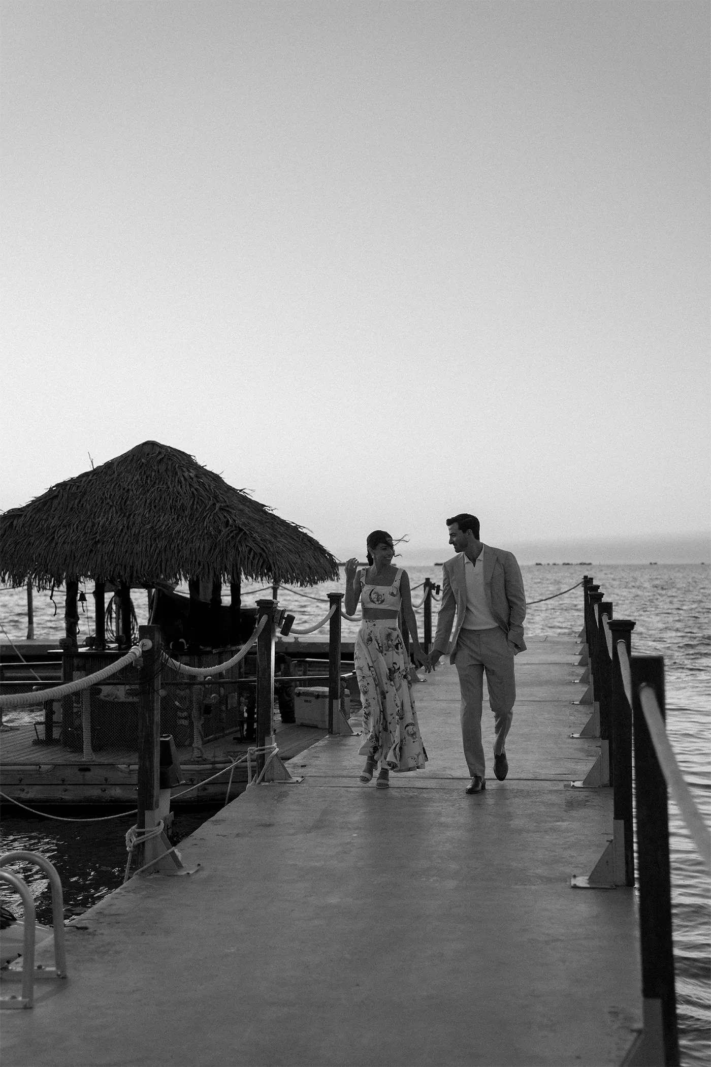 Couple holding hands walking along a wooden dock past a thatched hut at dusk