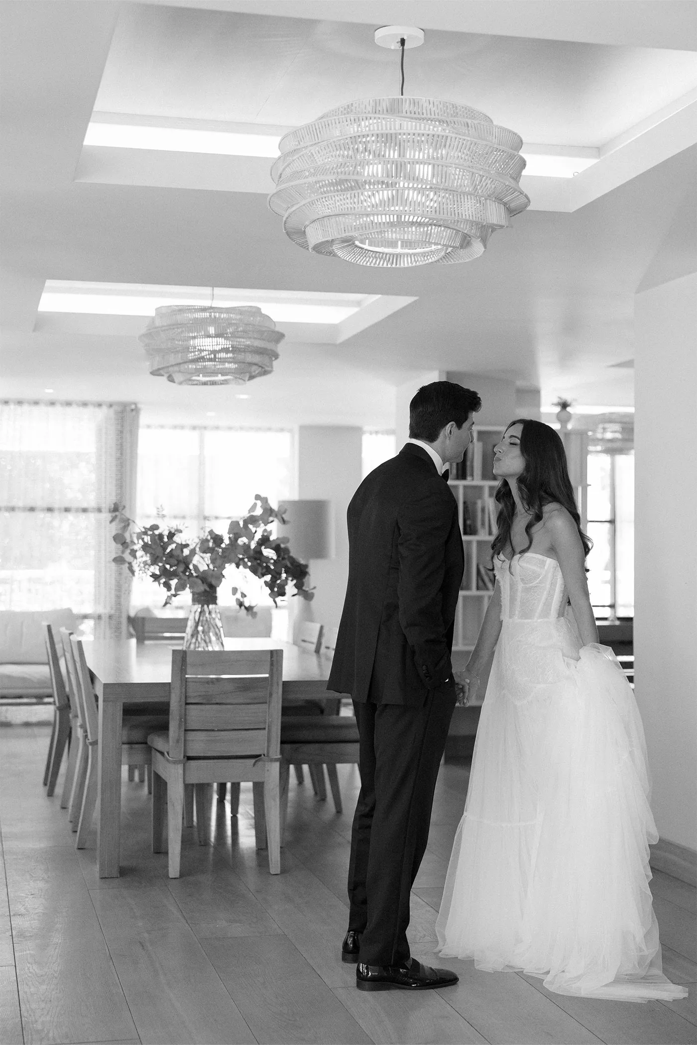 Bride and groom holding hands and leaning in to kiss in a bright dining room