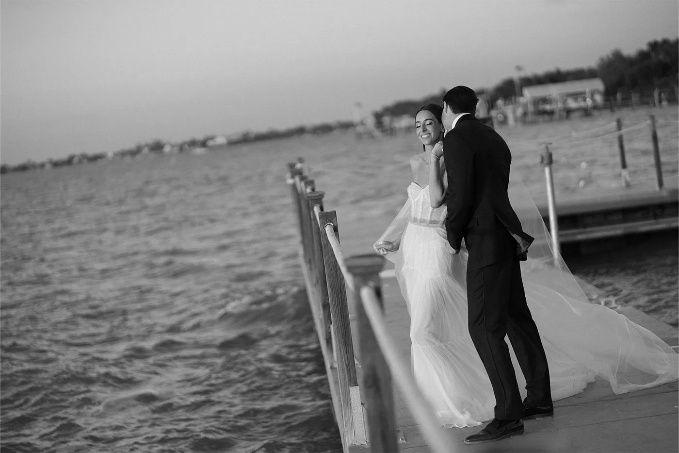 Bride and groom on a wooden dock at dusk, her veil trailing behind them over the water