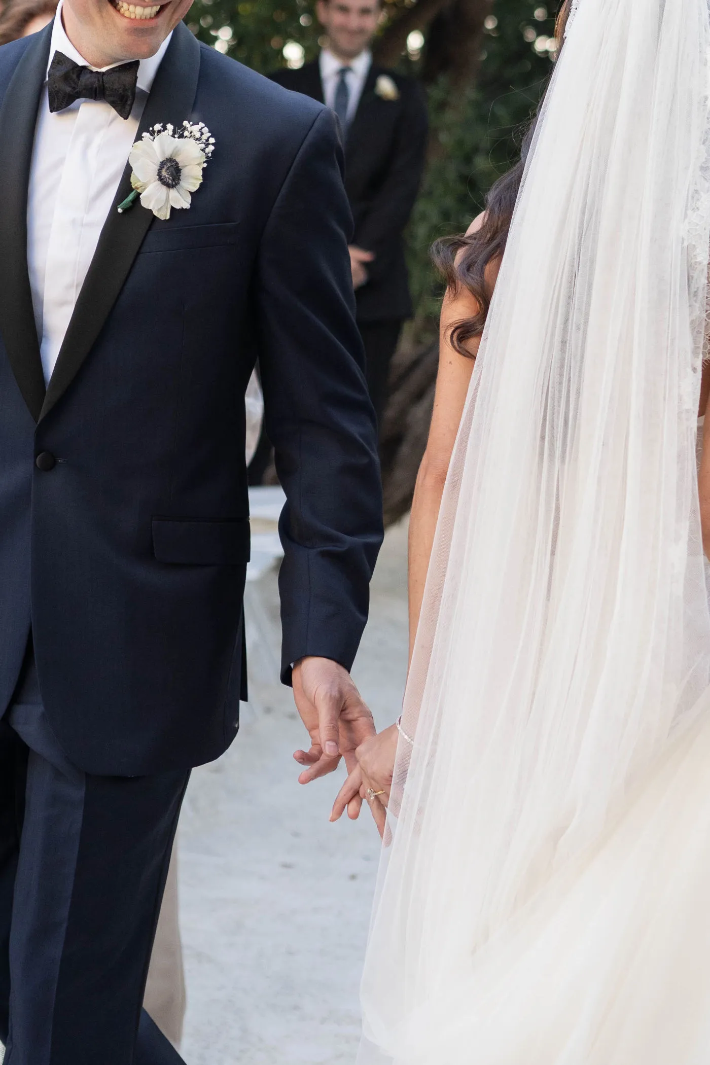 Close view of a groom in a tuxedo reaching for the bride's hand, veil beside him