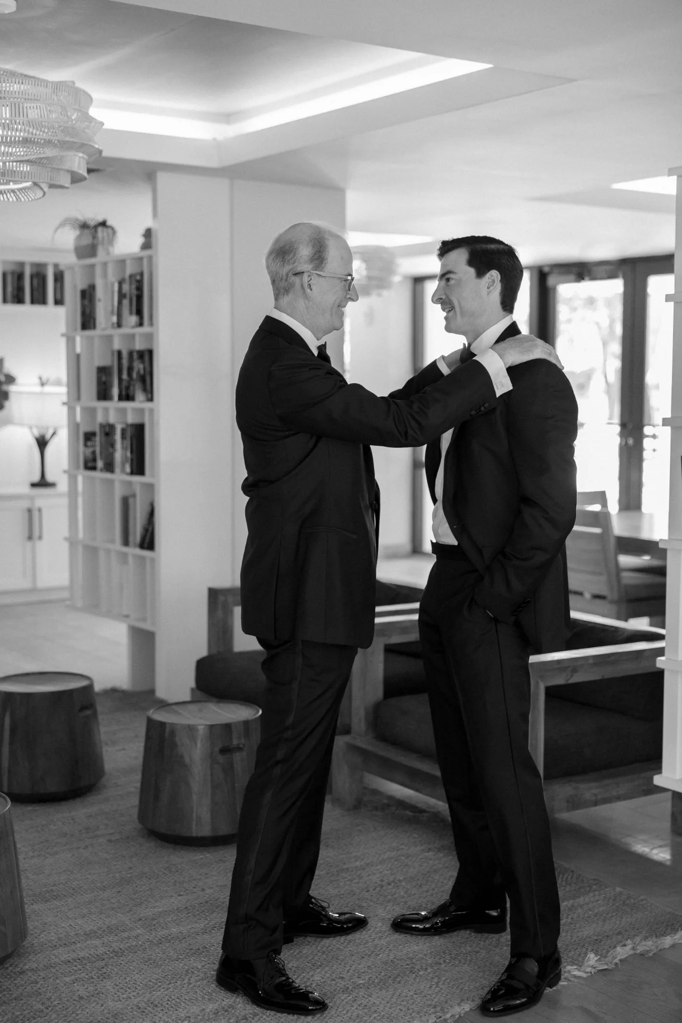 Older man straightening the groom's tuxedo jacket in a sunlit living room