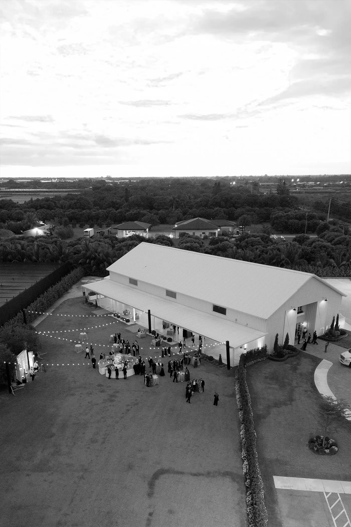 Aerial view of guests at a cocktail hour outside a white barn venue at dusk