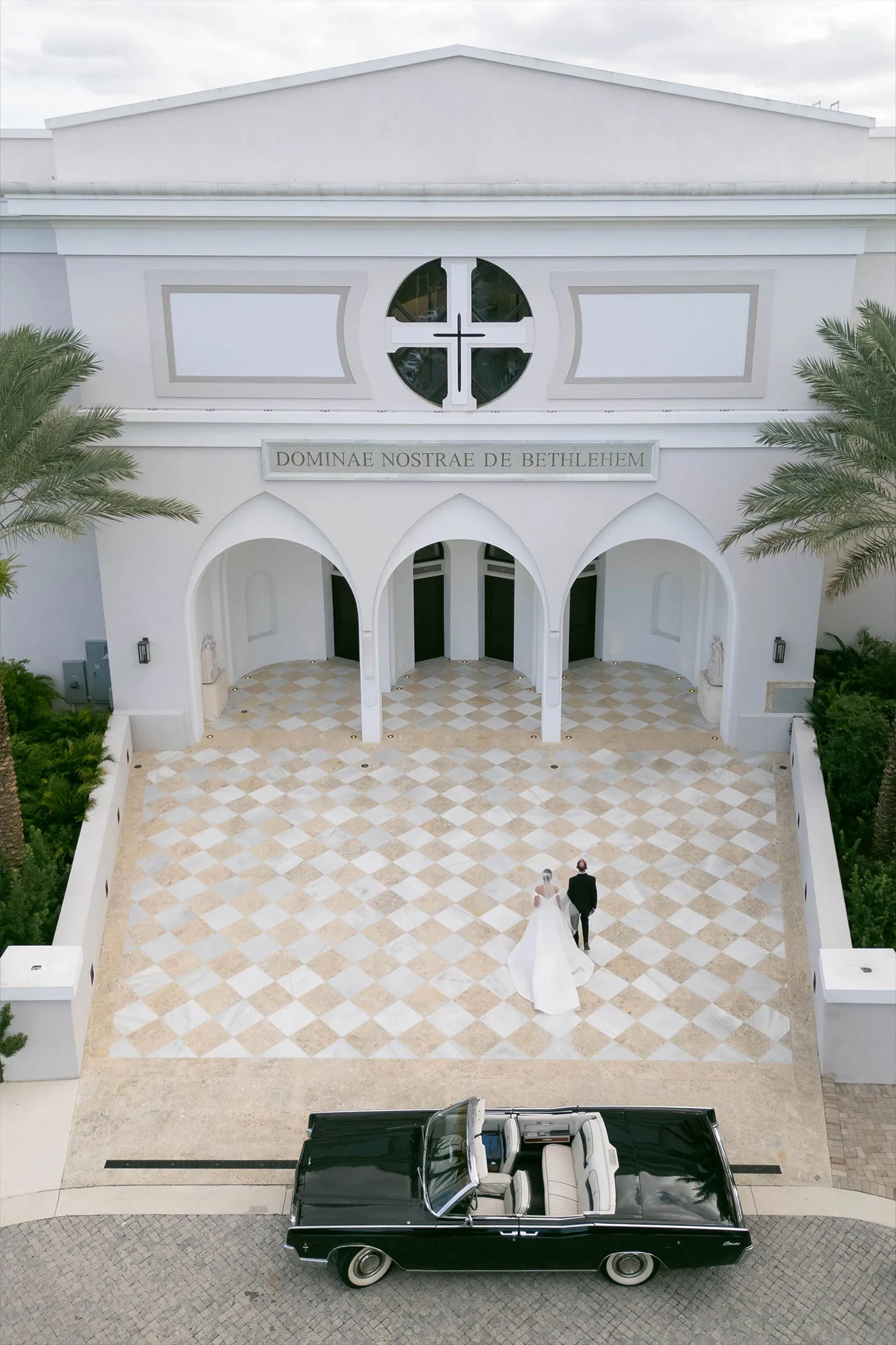Aerial view of a couple crossing a checkered church courtyard, vintage convertible waiting below