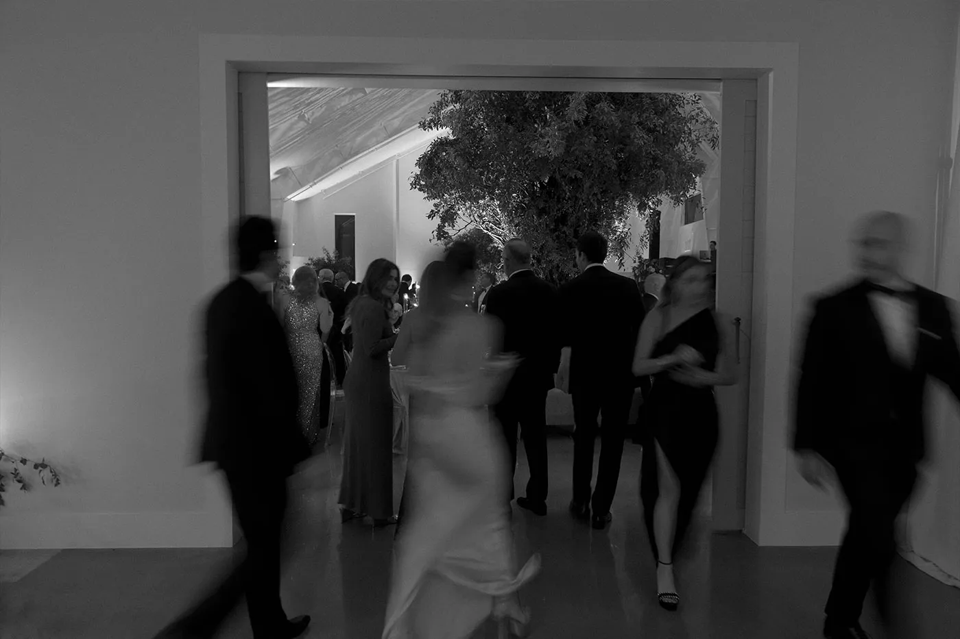 Guests in black tie blurred in motion through a doorway into a tree-filled reception
