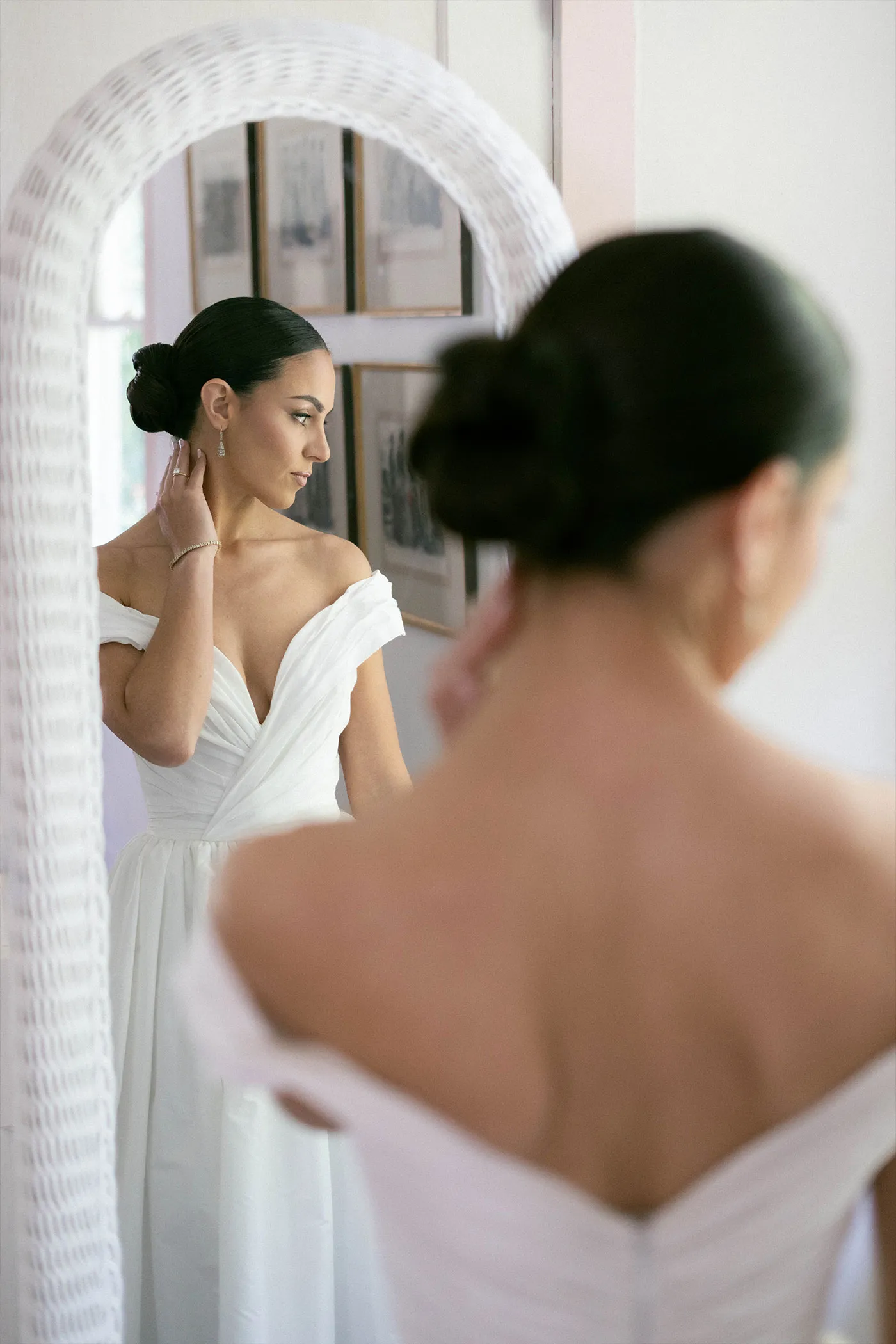 Bride in an off-shoulder gown fastening an earring in front of a mirror