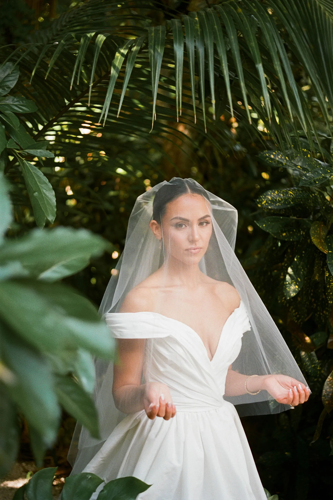 Bride in an off-shoulder gown and veil standing among palm fronds and tropical leaves