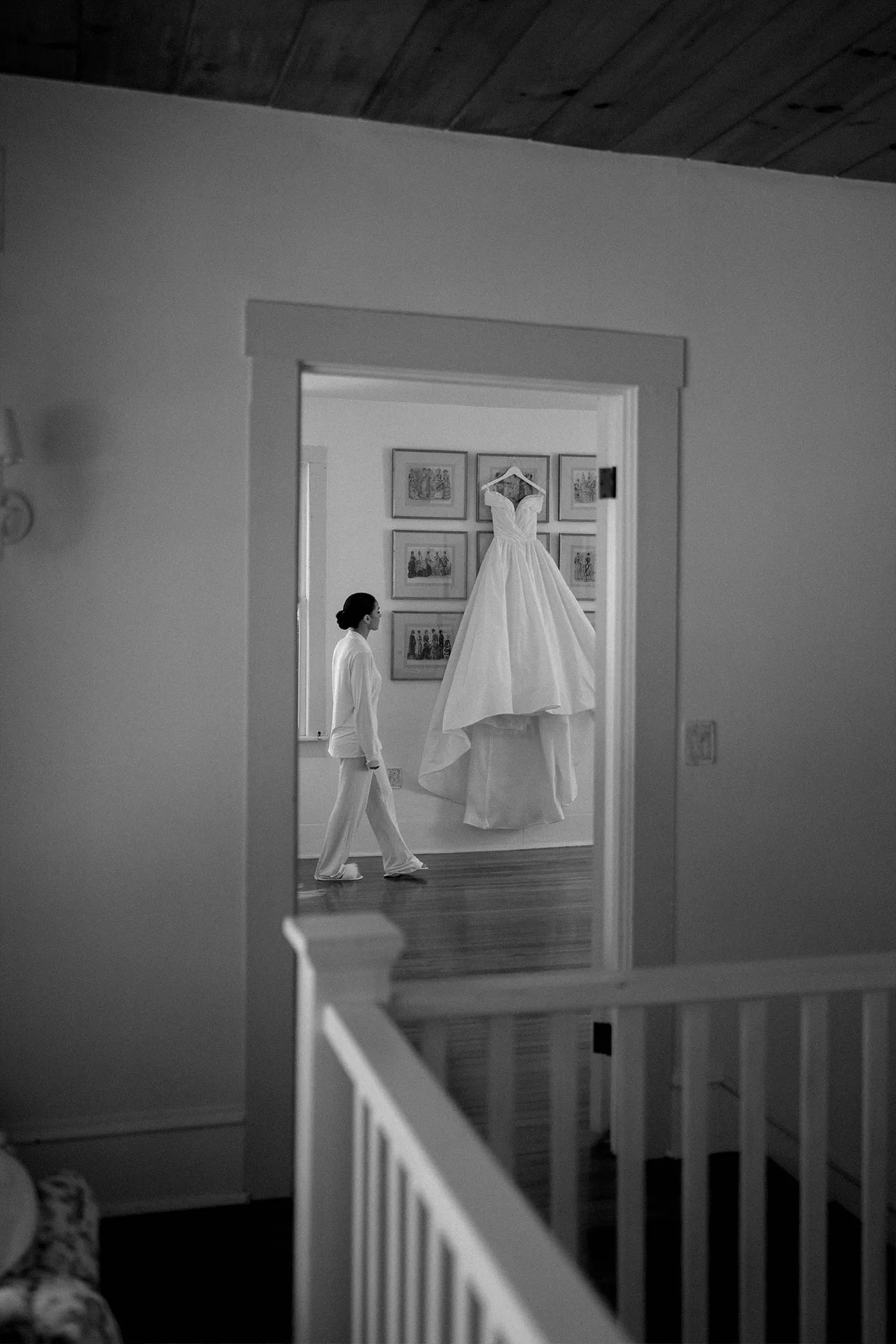 Bride walking past her gown hanging on the wall, seen through a doorway