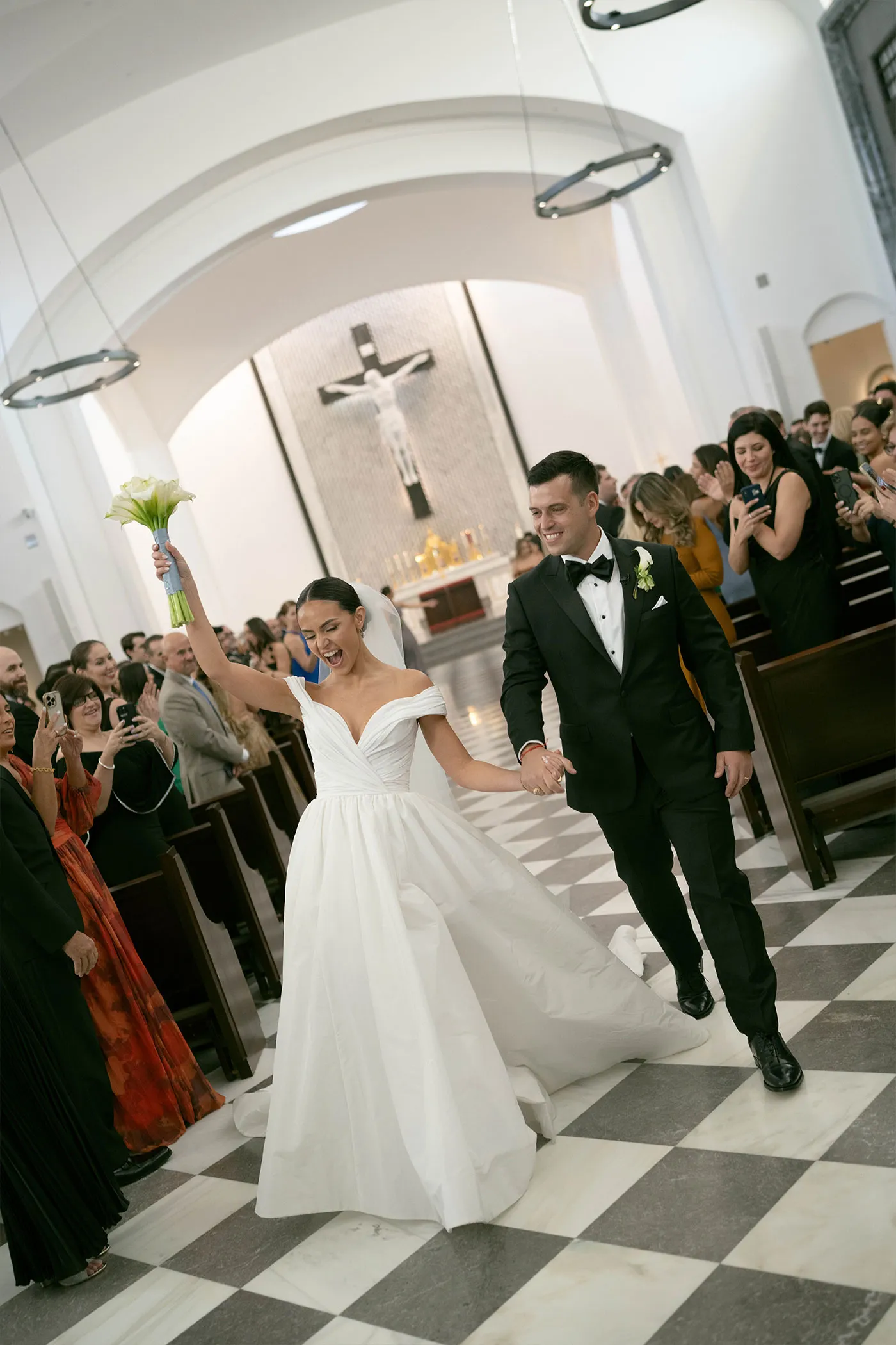 Bride raising her bouquet as she and the groom walk back up the aisle past clapping guests