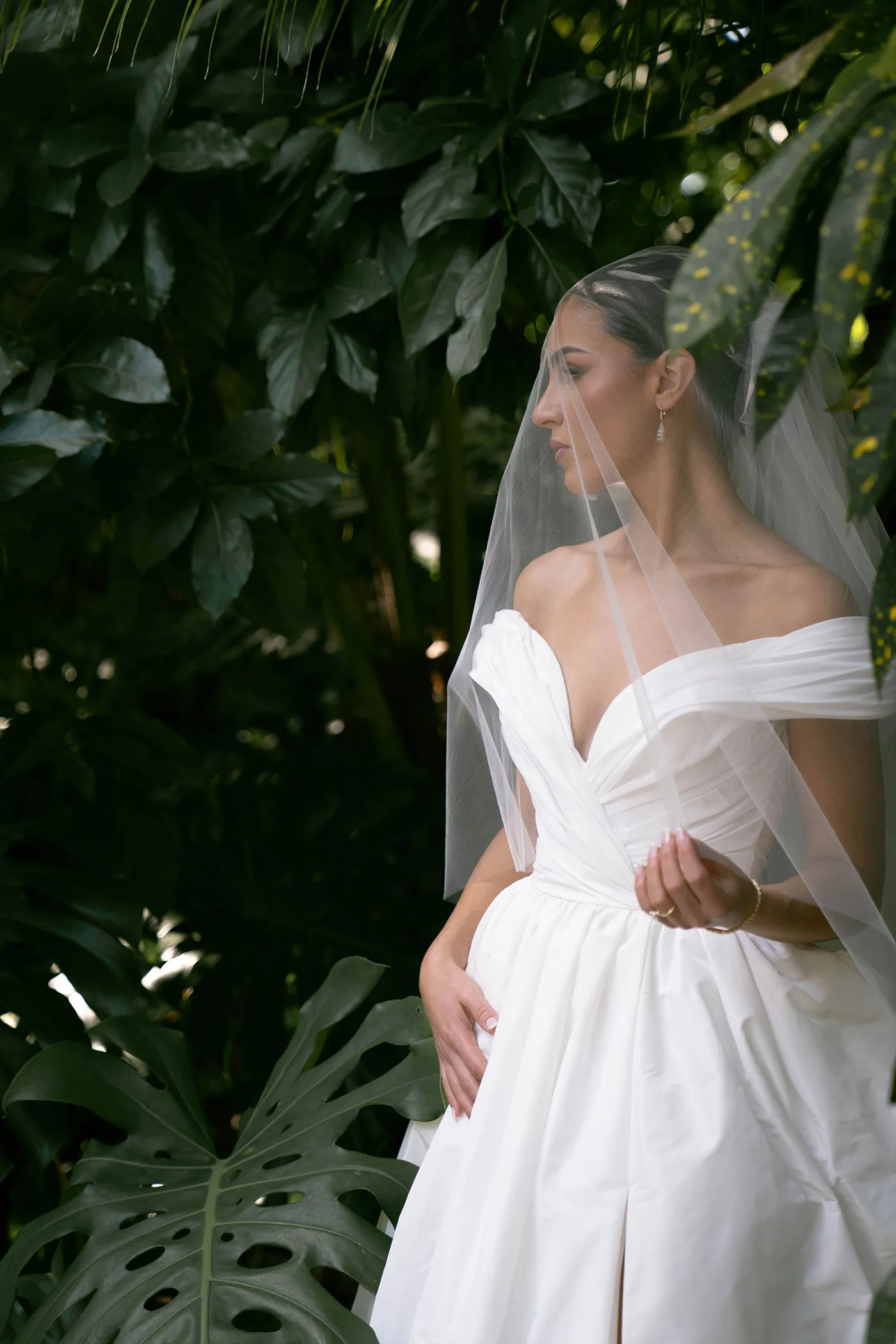 Bride in a veil and off-shoulder gown standing among large tropical leaves