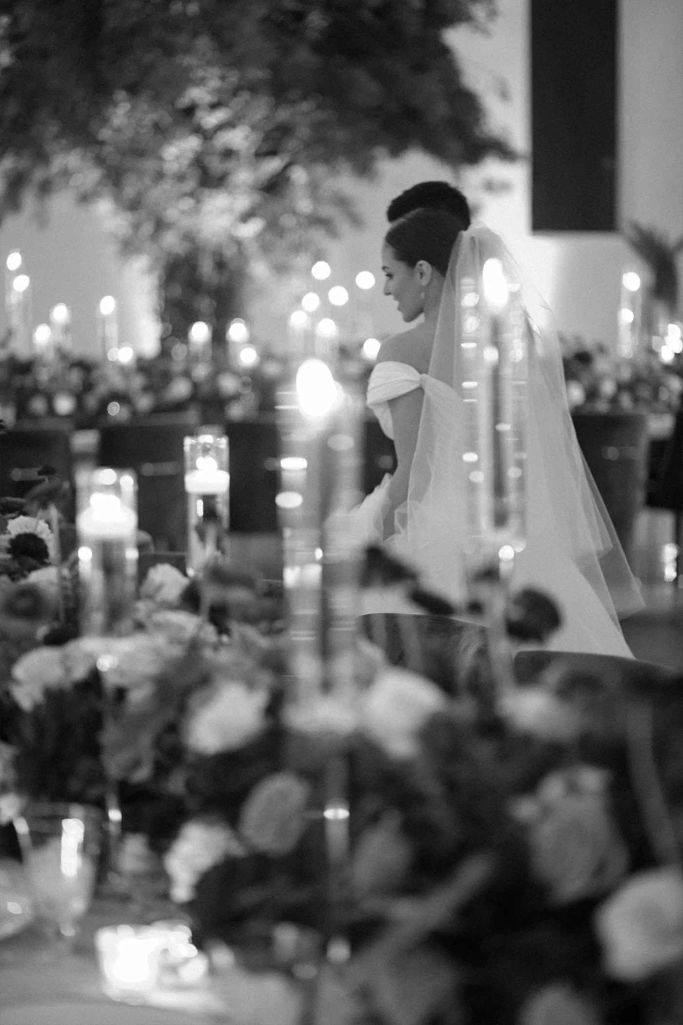 Bride in a veil seen past candles and flowers at a reception table, black and white