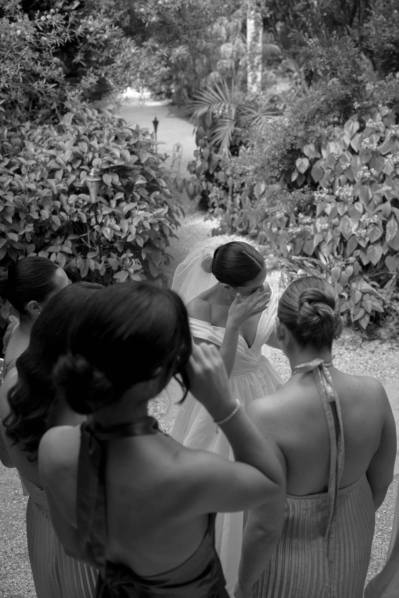 Bridesmaids fixing the bride's veil on a garden path, seen from behind in black and white
