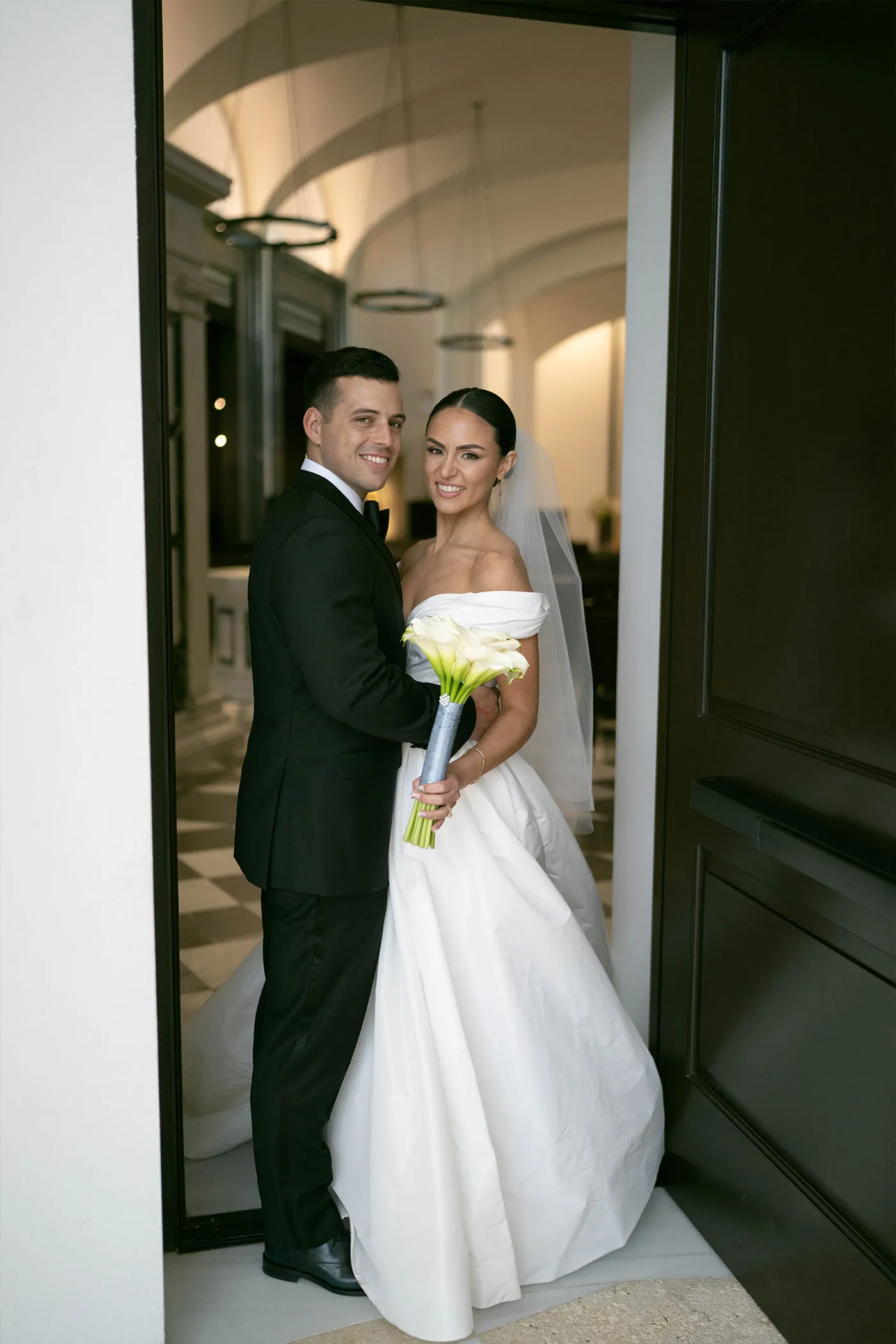 Bride with a calla lily bouquet and groom in a tuxedo standing in a dark doorway