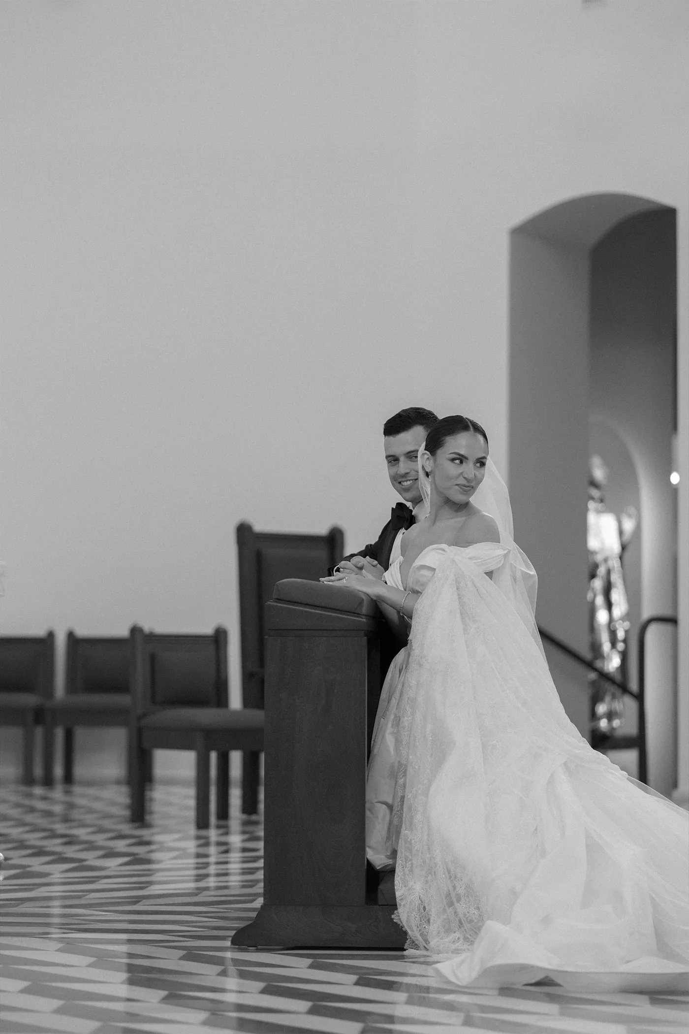Bride and groom kneeling at a prayer bench in a church, her train spread behind, black and white