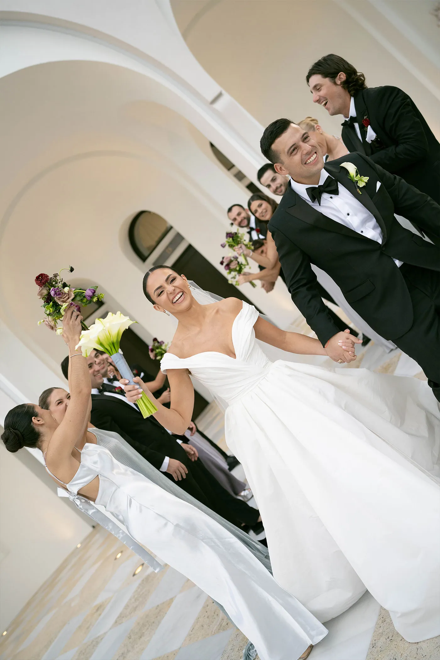 Laughing bride and groom leading the wedding party down steps, bouquets raised
