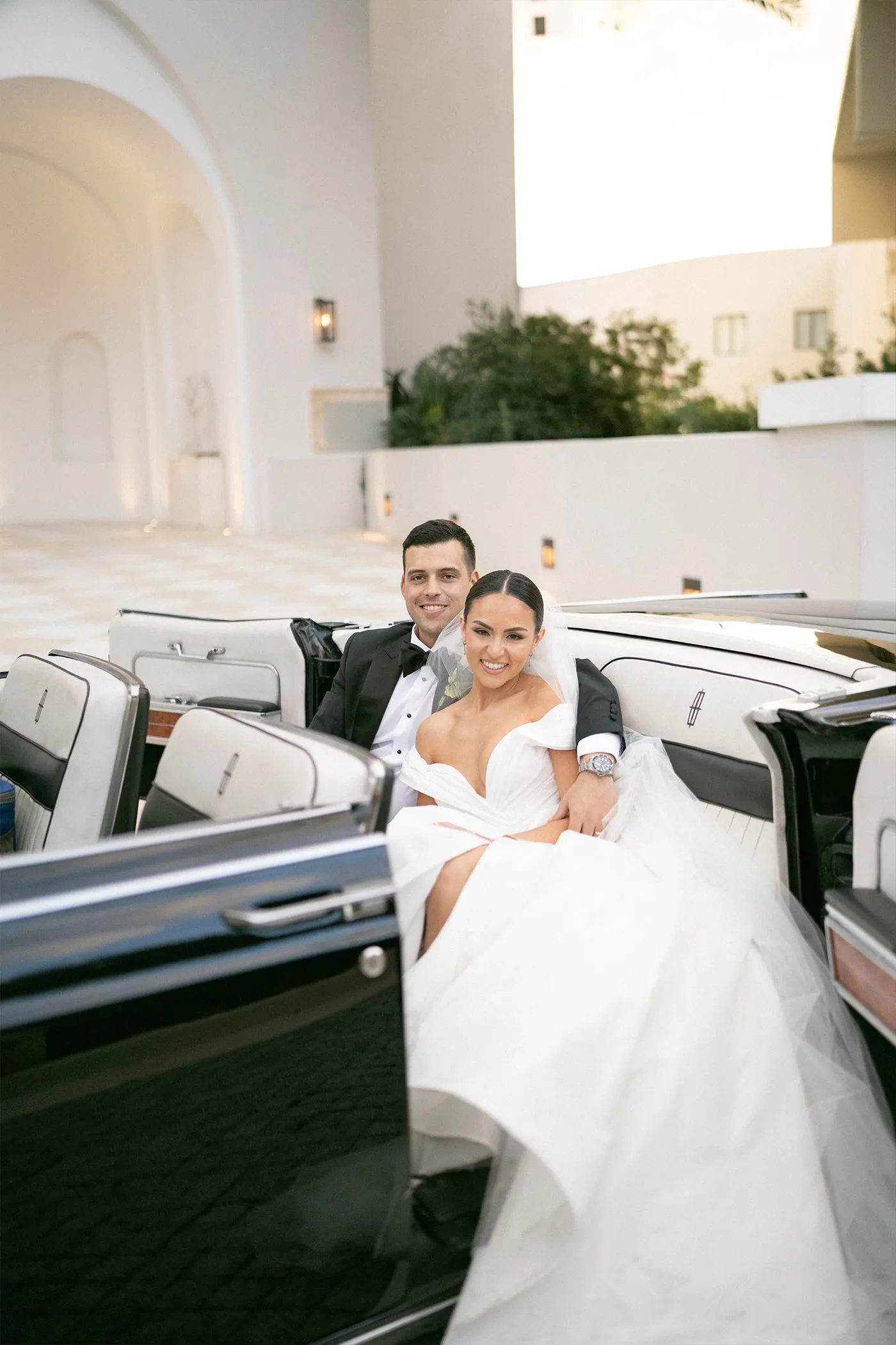 Bride and groom seated in the back of a vintage convertible outside a white venue