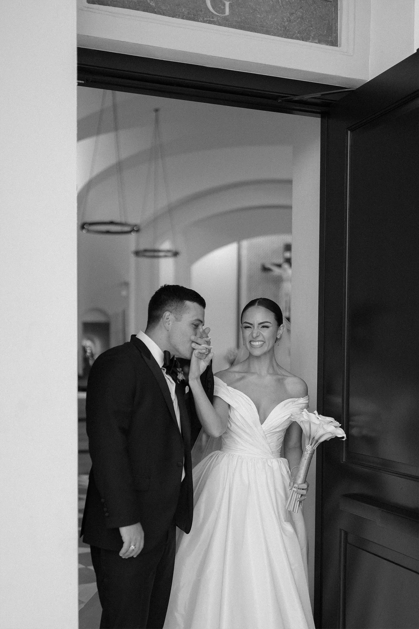Groom kissing the laughing bride's hand in a doorway, black and white