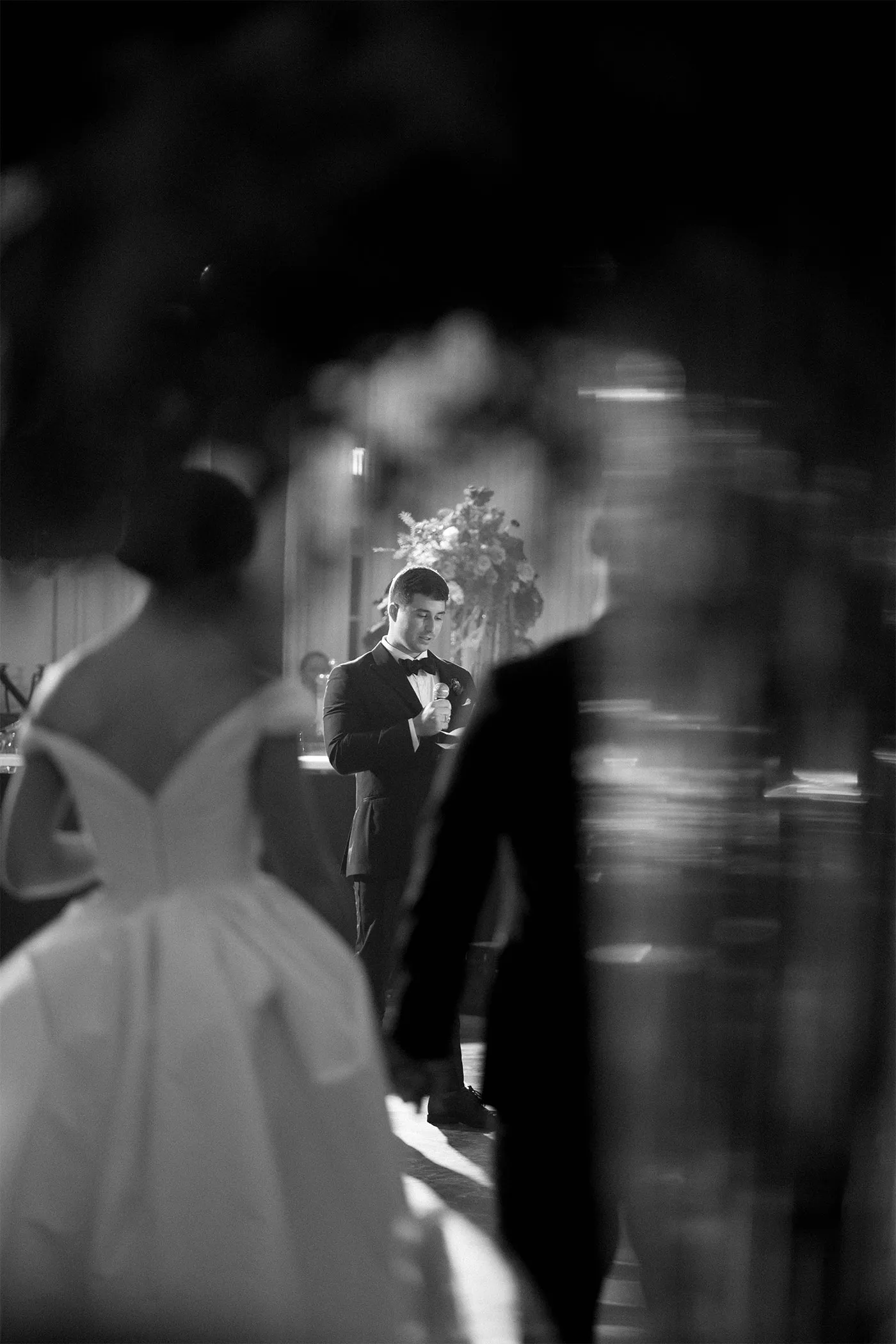 Groom in a tuxedo giving a speech, framed between blurred guests