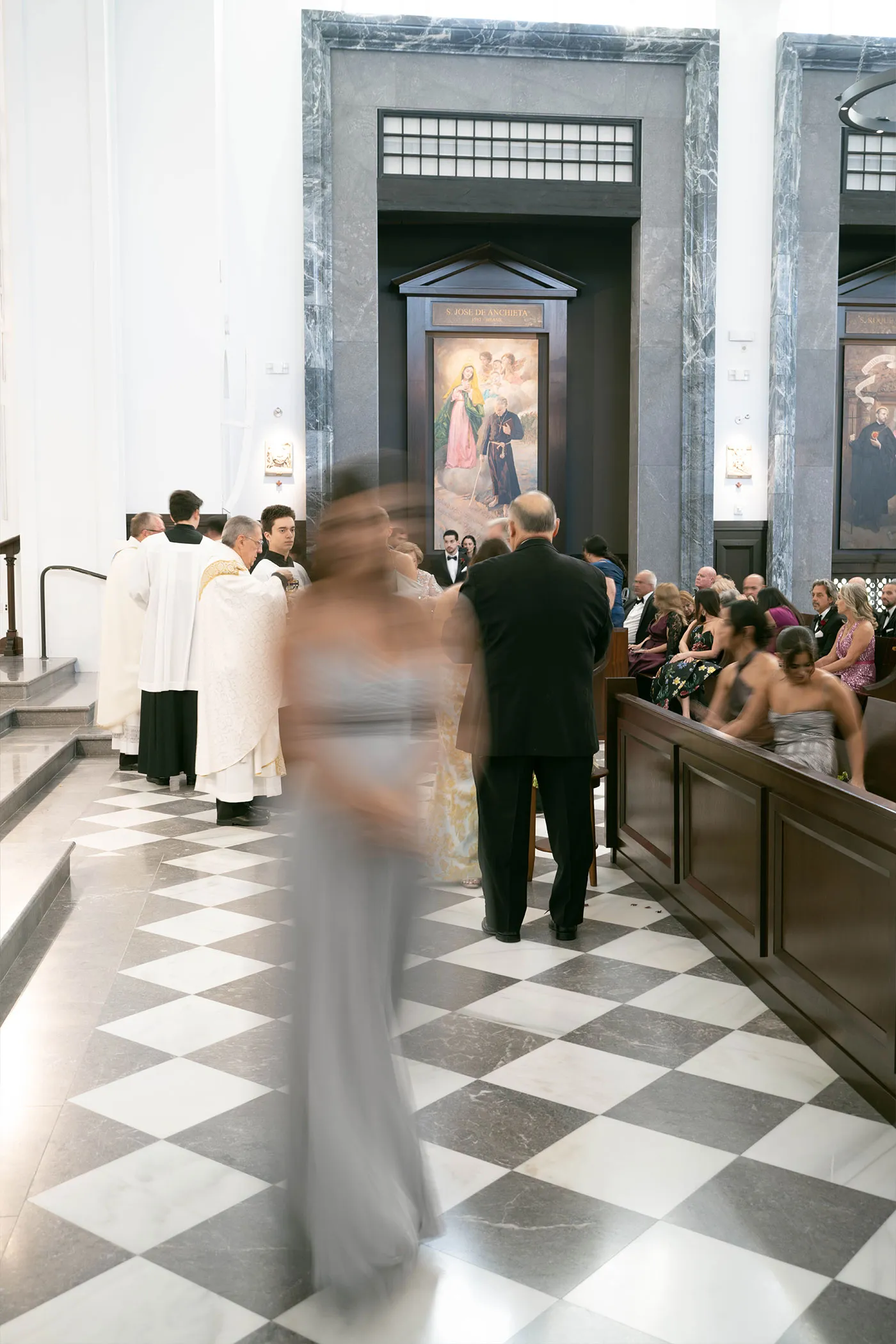 Guest in a grey gown blurred in motion crossing a checkered church aisle past clergy