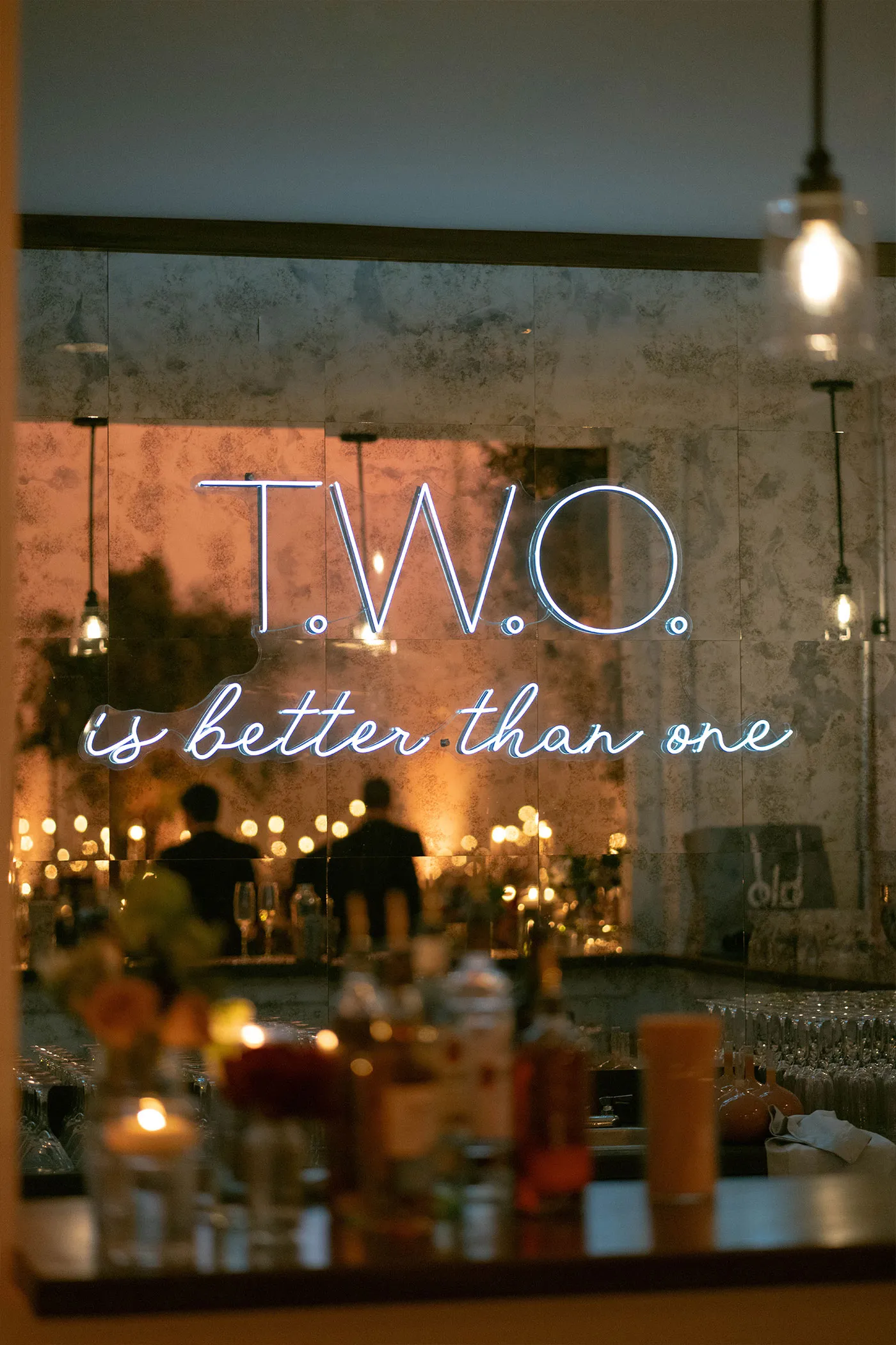 Neon sign reading T.W.O. is better than one above a candlelit reception bar