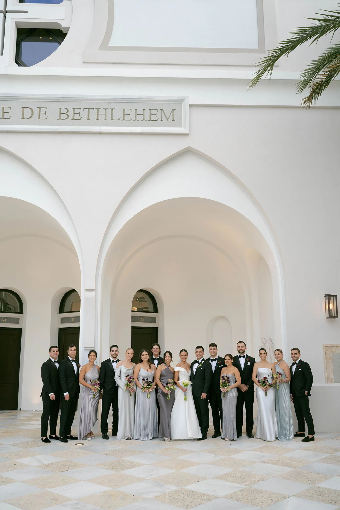 Wedding party in tuxedos and silver gowns outside a white arched church entrance