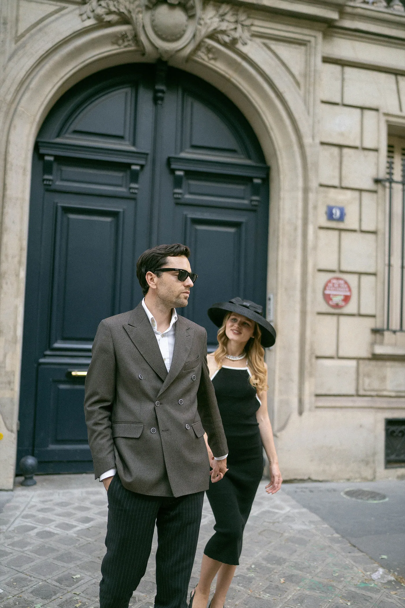 Couple in tailored suit and black hat walking past an arched navy door