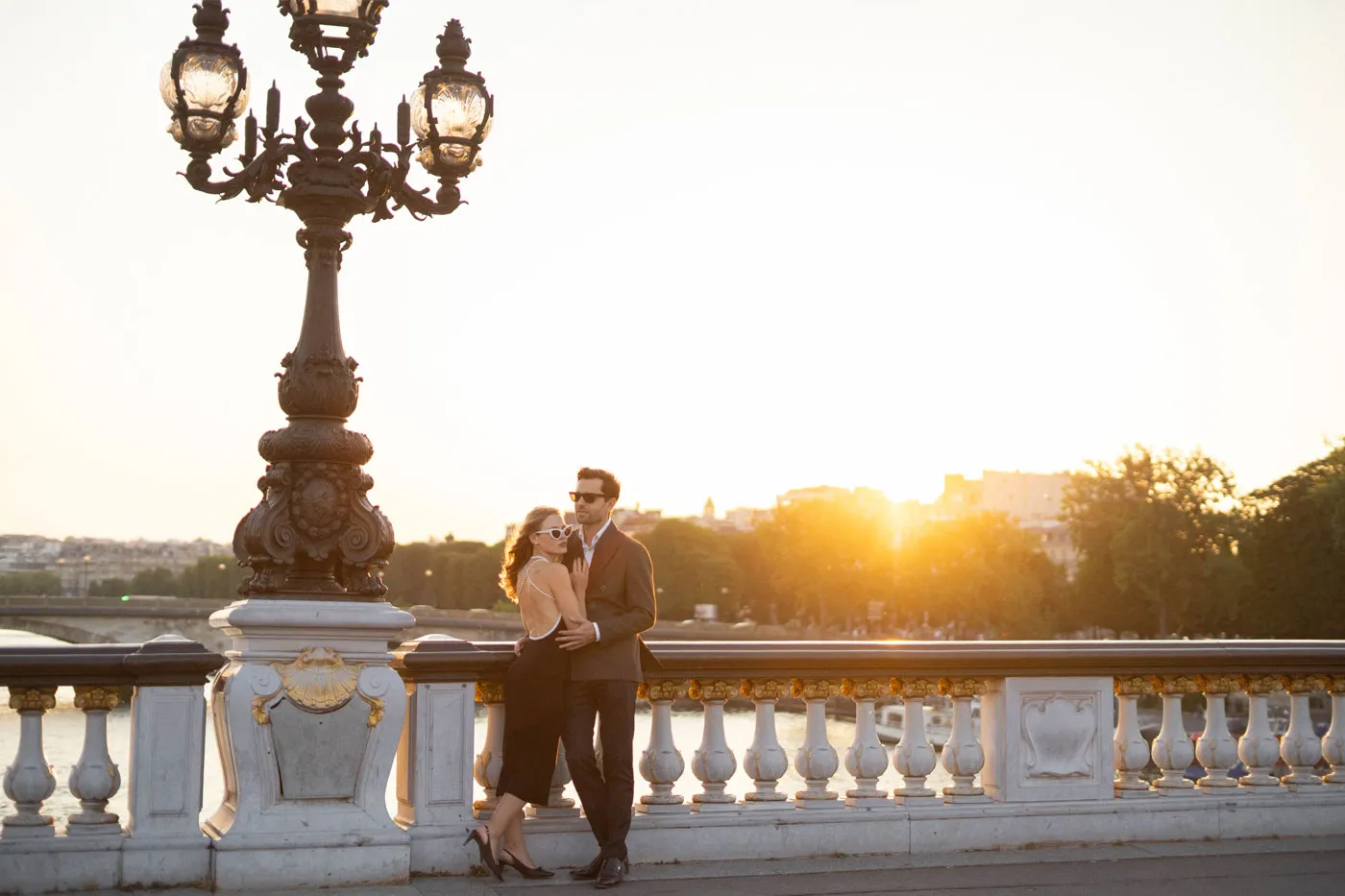 Couple embracing at an ornate bridge railing under a lamppost at sunset
