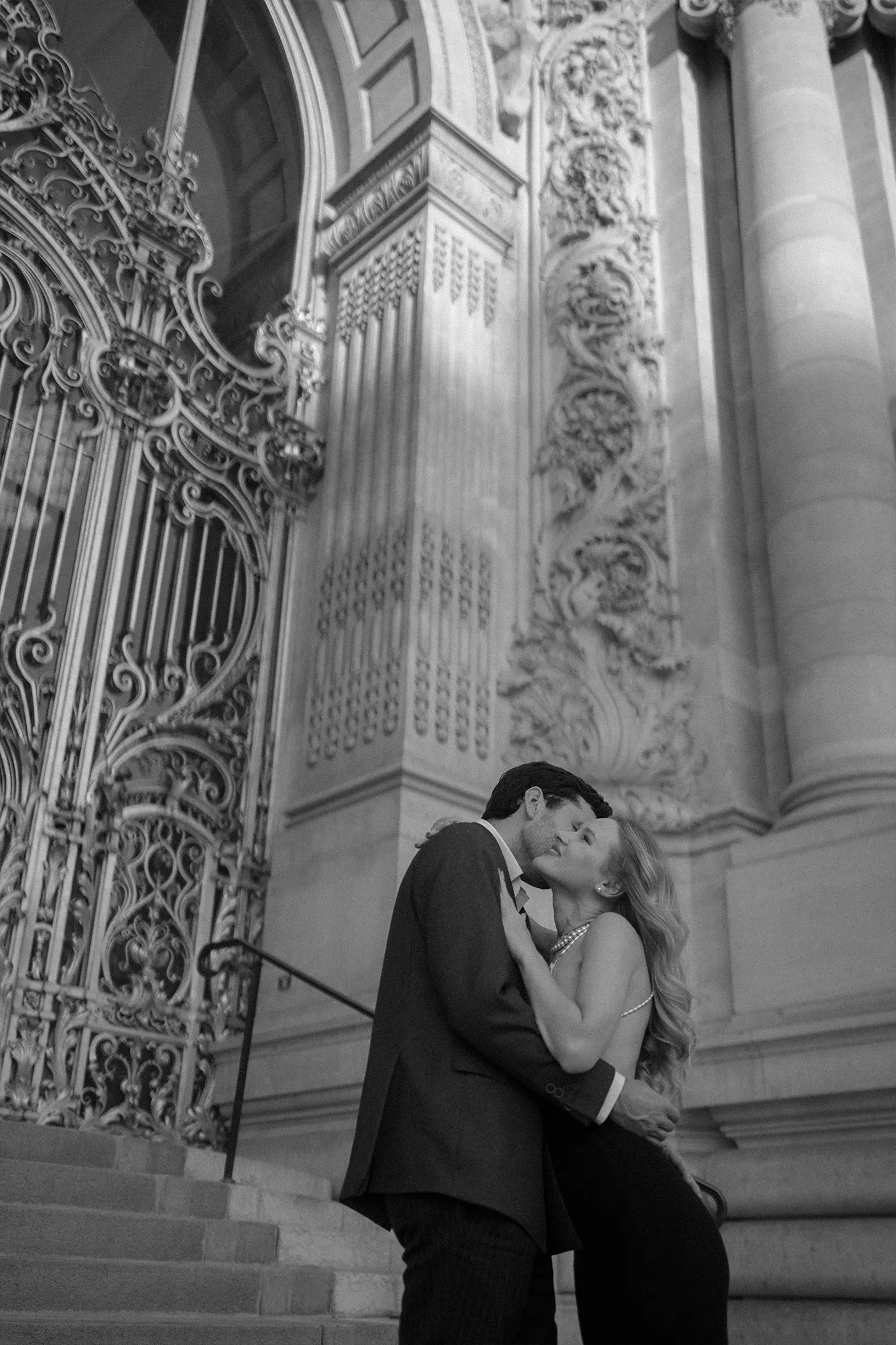 Couple kissing on the steps of an ornate stone building with carved ironwork doors